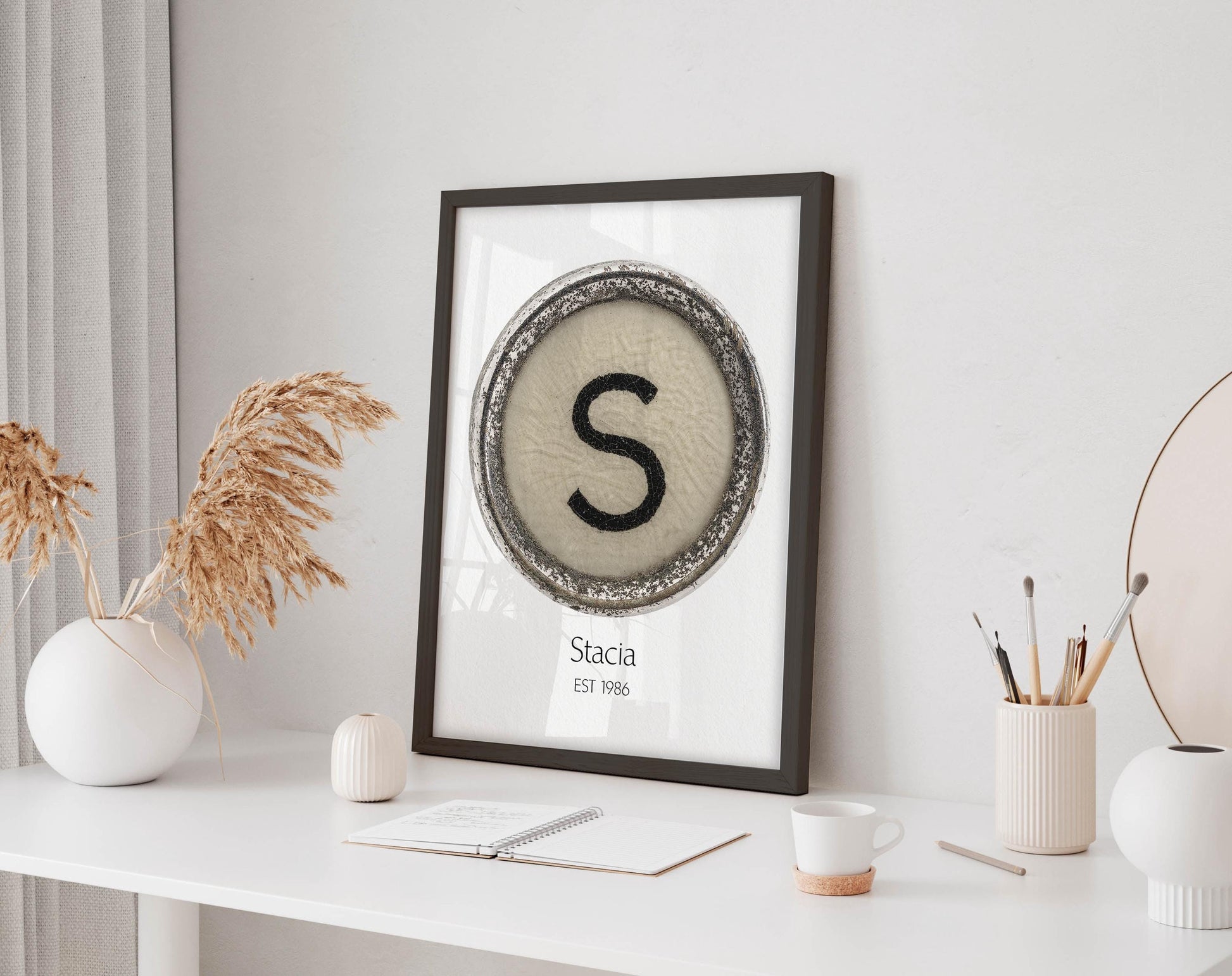 a framed artwork with a large letter 'S' on a white background, placed on a white desk. The desk also has a notebook, a cup, and a vase with dried flowers.