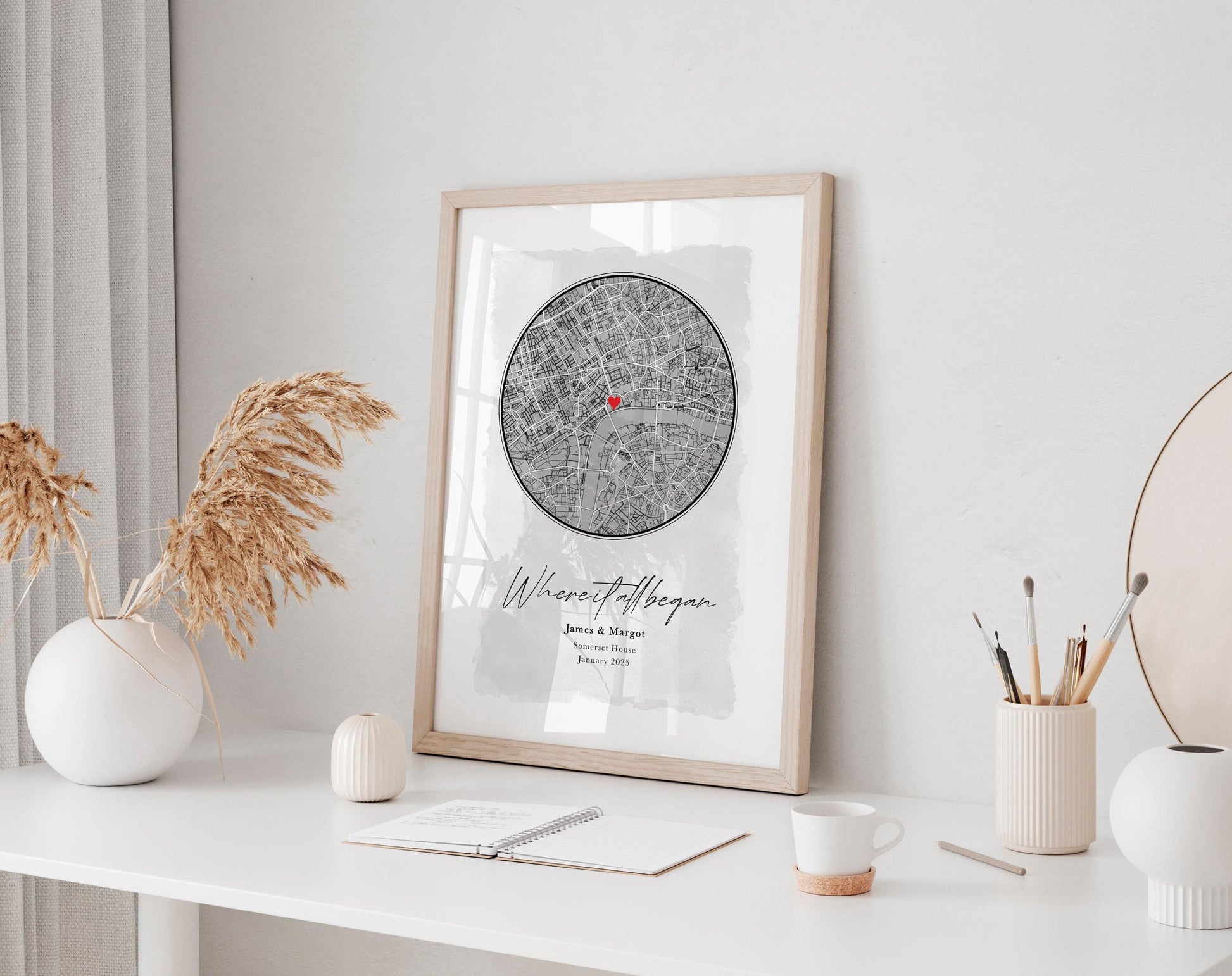 a framed map of London with a decorative border, placed on a white desk. The desk also has a notebook, a cup, and a vase with dried flowers.