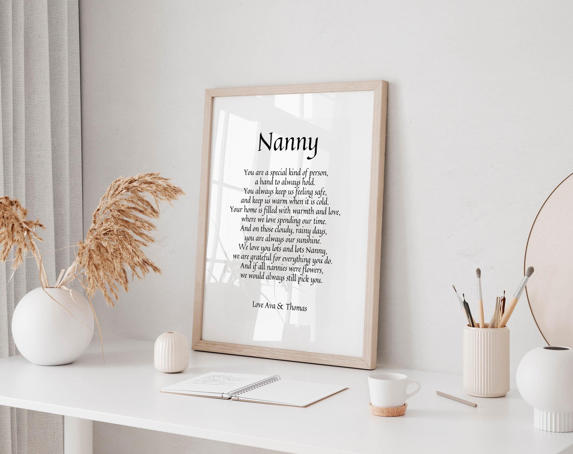 a framed quote by Ava Thomas titled "Nanny" on a white wall, accompanied by a small notebook, a white mug, and a white vase with dried flowers.
