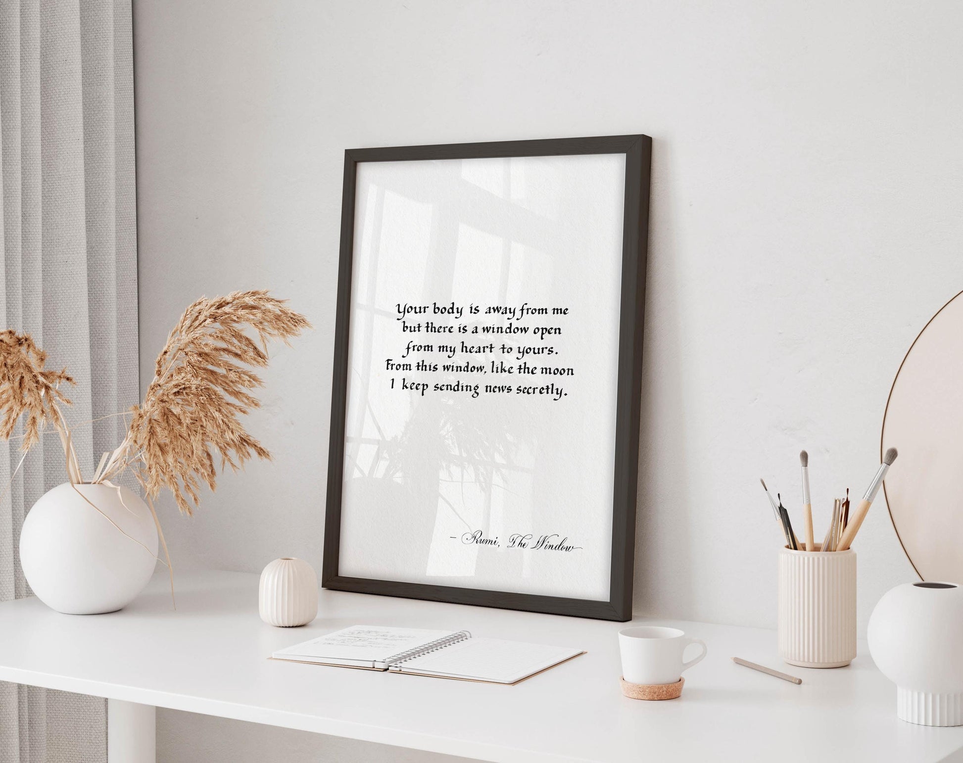 a framed quote on a white desk, surrounded by various objects such as a notebook, a cup, and a vase with dried flowers.