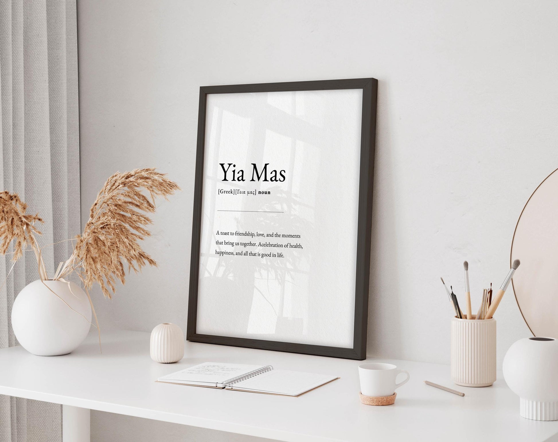 a framed definition of 'Yia Mas' on a white desk, accompanied by a notebook, a cup, and a vase with dried flowers.