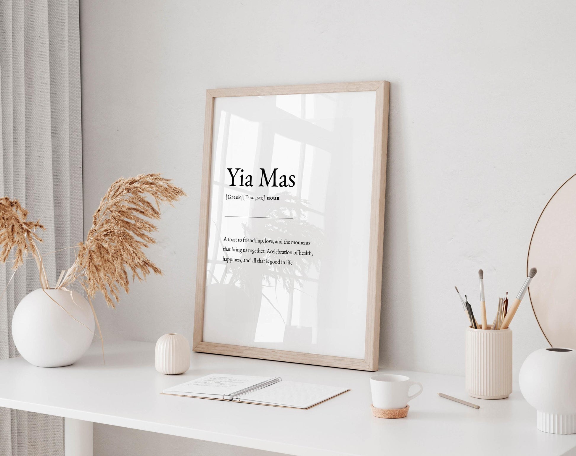 a minimalist workspace with a framed definition of 'Yia Mas' on a white wall, a white desk with a notebook, a white mug, and a white vase with dried flowers.