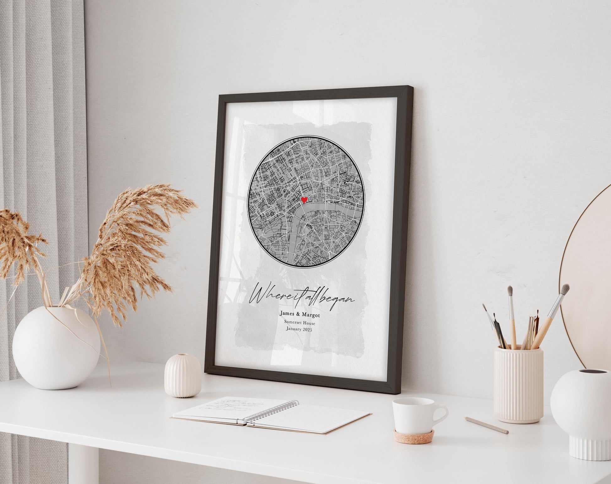 a framed map of London with a personal touch, displaying the name 'James & Margaret' and the date 'January 2015'. The map is hung on a white wall, and there is a desk with various items on it, including a notebook, a cup, and a vase with dried flowers.