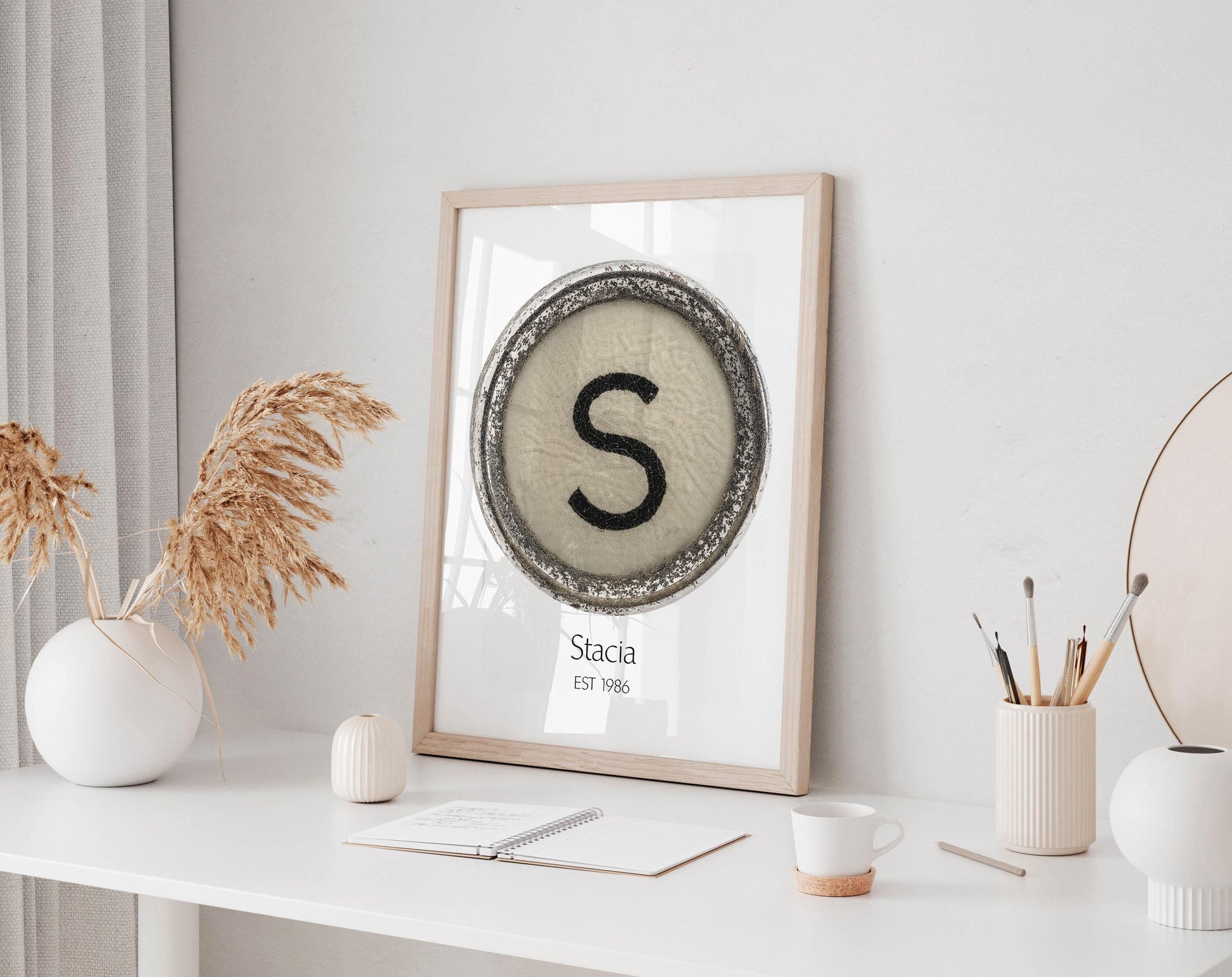 a framed poster with a large letter 'S' on it, placed on a white desk. The desk also has a notebook, a cup, and a vase with dried flowers.