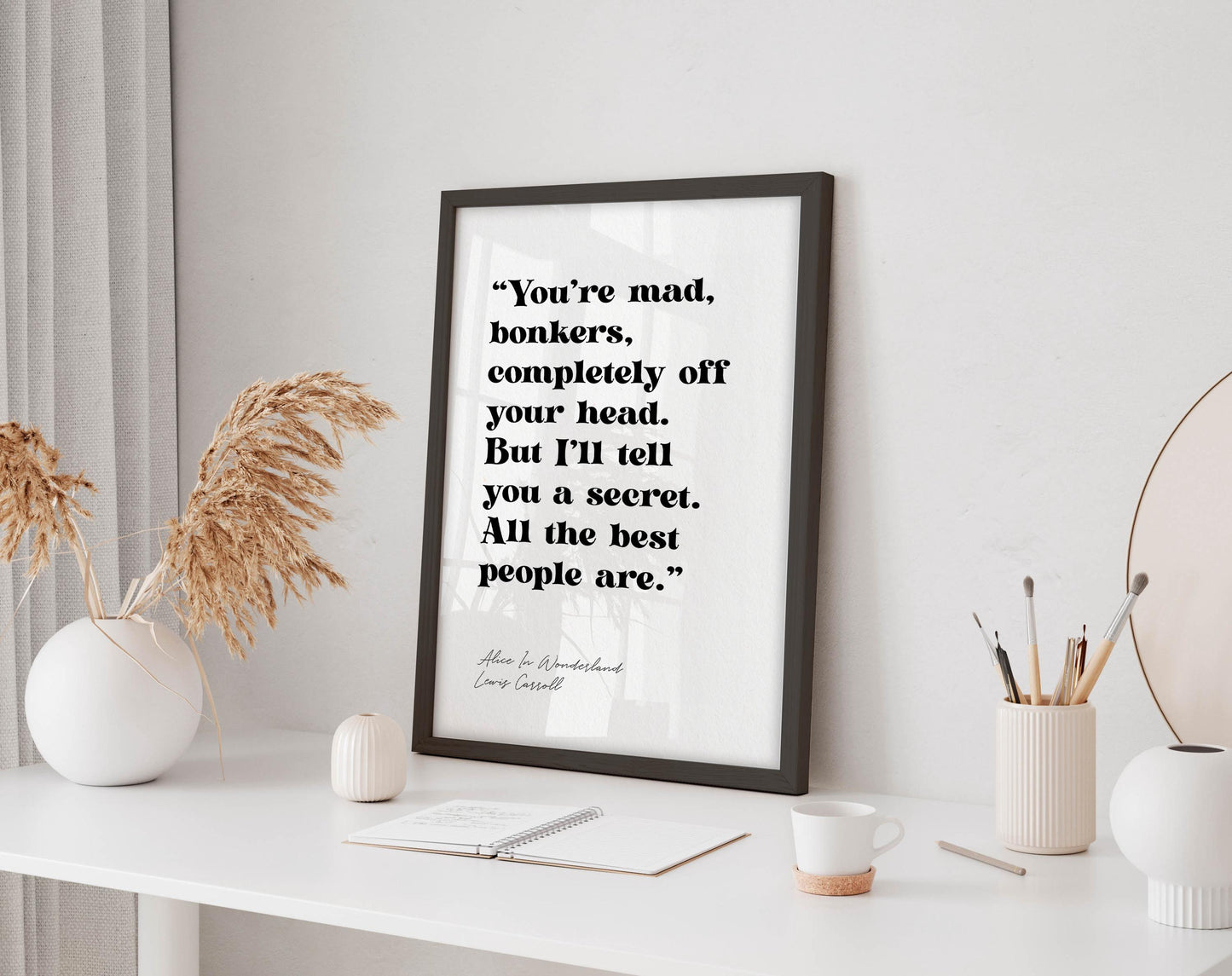 a framed quote on a white desk, surrounded by various objects such as a notebook, a cup, and a vase with dried flowers.