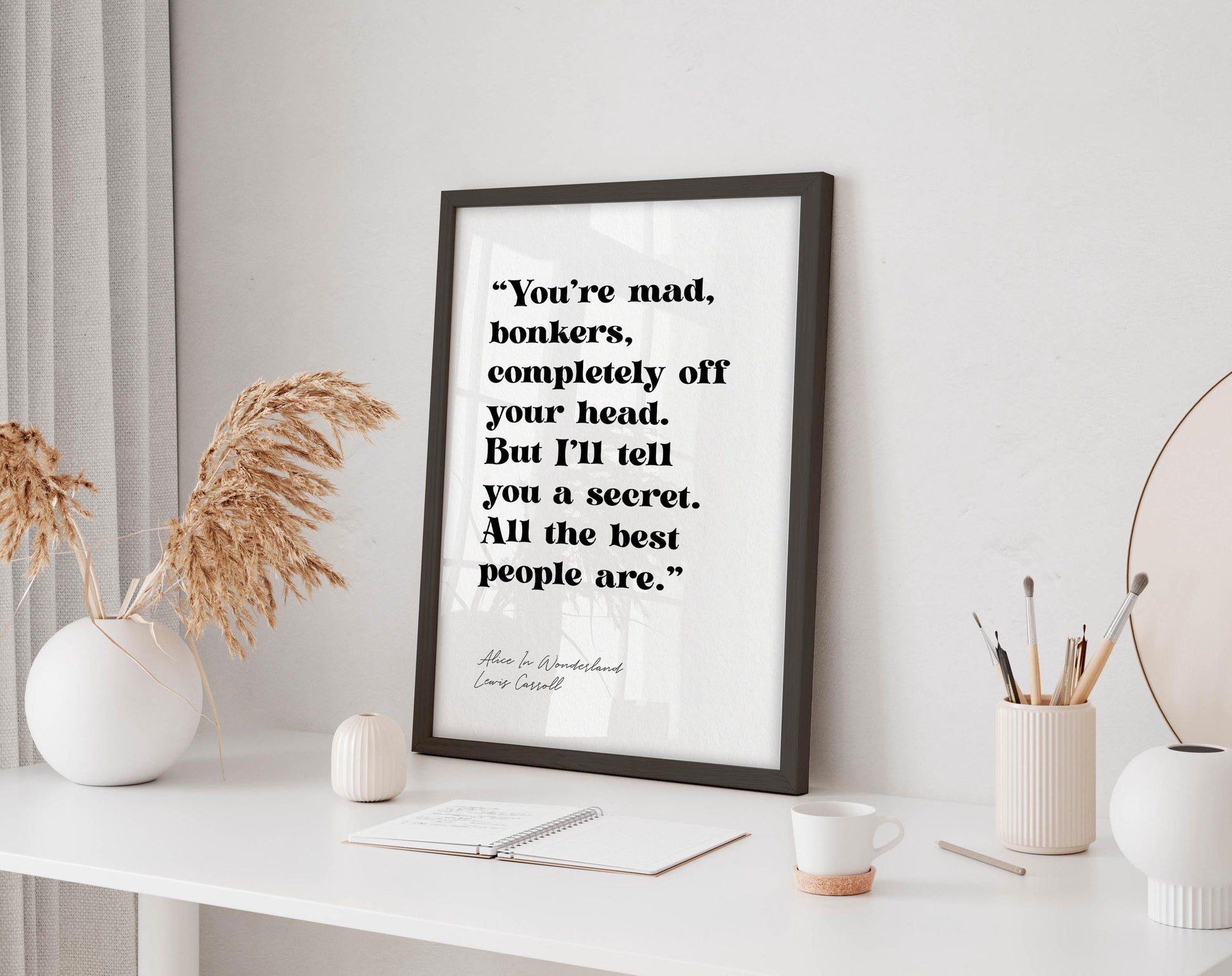 a framed quote on a white desk, surrounded by various objects such as a notebook, a cup, and a vase with dried flowers.