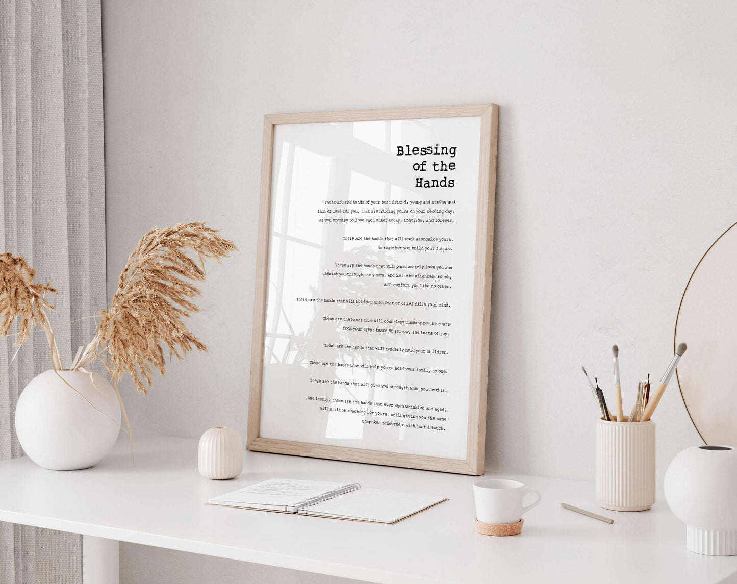 A framed print titled &quot;Blessing of the Hands&quot; is displayed on a white desk, accompanied by a notebook, a cup, and a vase with dried flowers.
