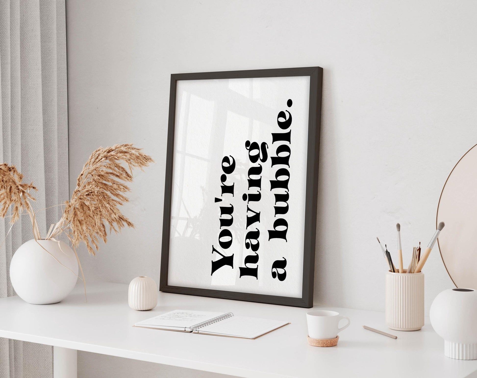 a white desk with a framed quote, a small white vase with dried flowers, a white mug, a small white candle, and a small white candle holder.