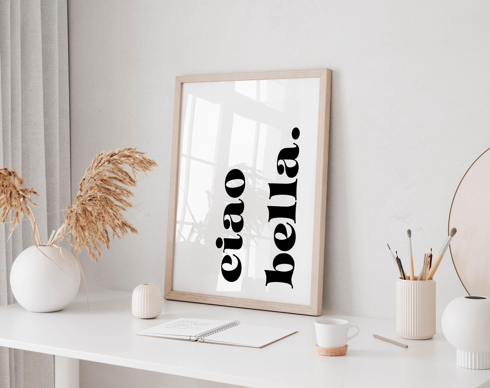a minimalist workspace with a white desk, a framed poster with the text &quot;ciao bella&quot; in black, a white vase with dried flowers, a white mug, and a small white figurine.