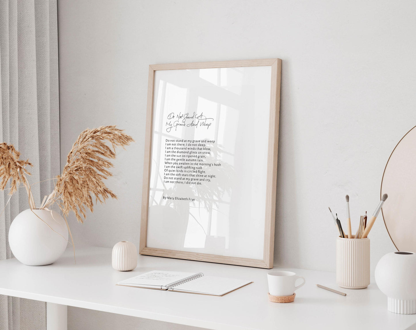 a white desk with a framed poem, a notebook, a cup, and a vase with dried flowers. The desk is set against a white wall, and there is a window in the background.