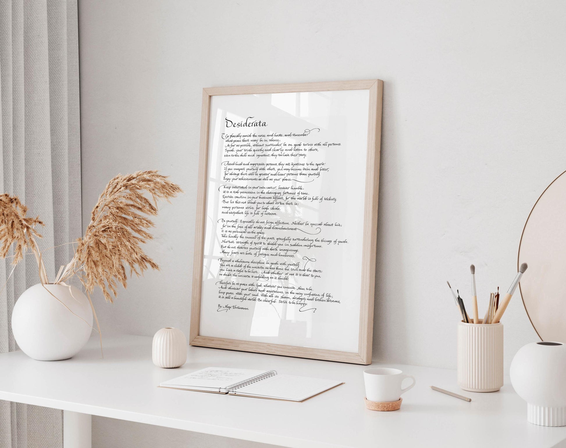 a framed poem titled &quot;Desiderata&quot; displayed on a white desk, accompanied by a notebook, a cup, and a vase with dried flowers.