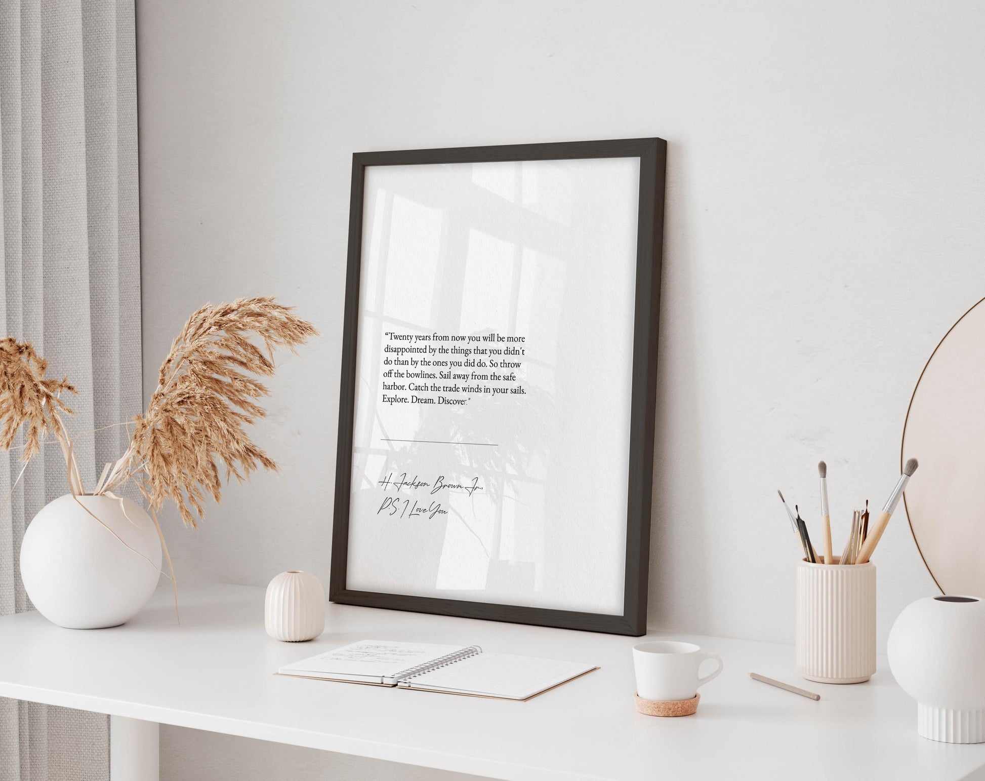 a white desk with a framed quote, a notebook, a cup, and a vase with dried flowers. The desk is set against a white wall, and there is a window in the background.
