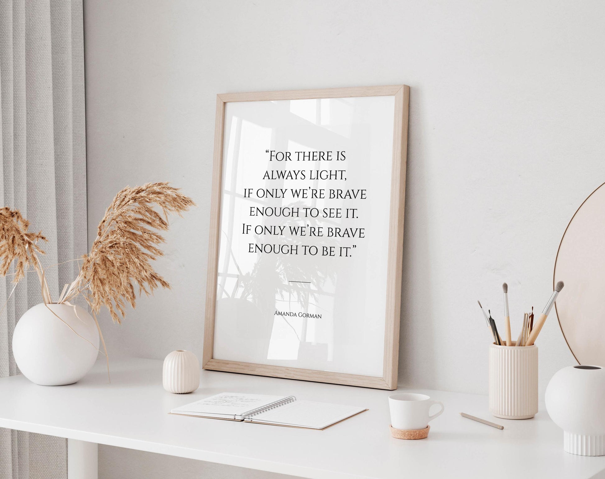 a framed quote on a white desk, surrounded by various objects such as a notebook, a cup, and a vase with dried flowers.