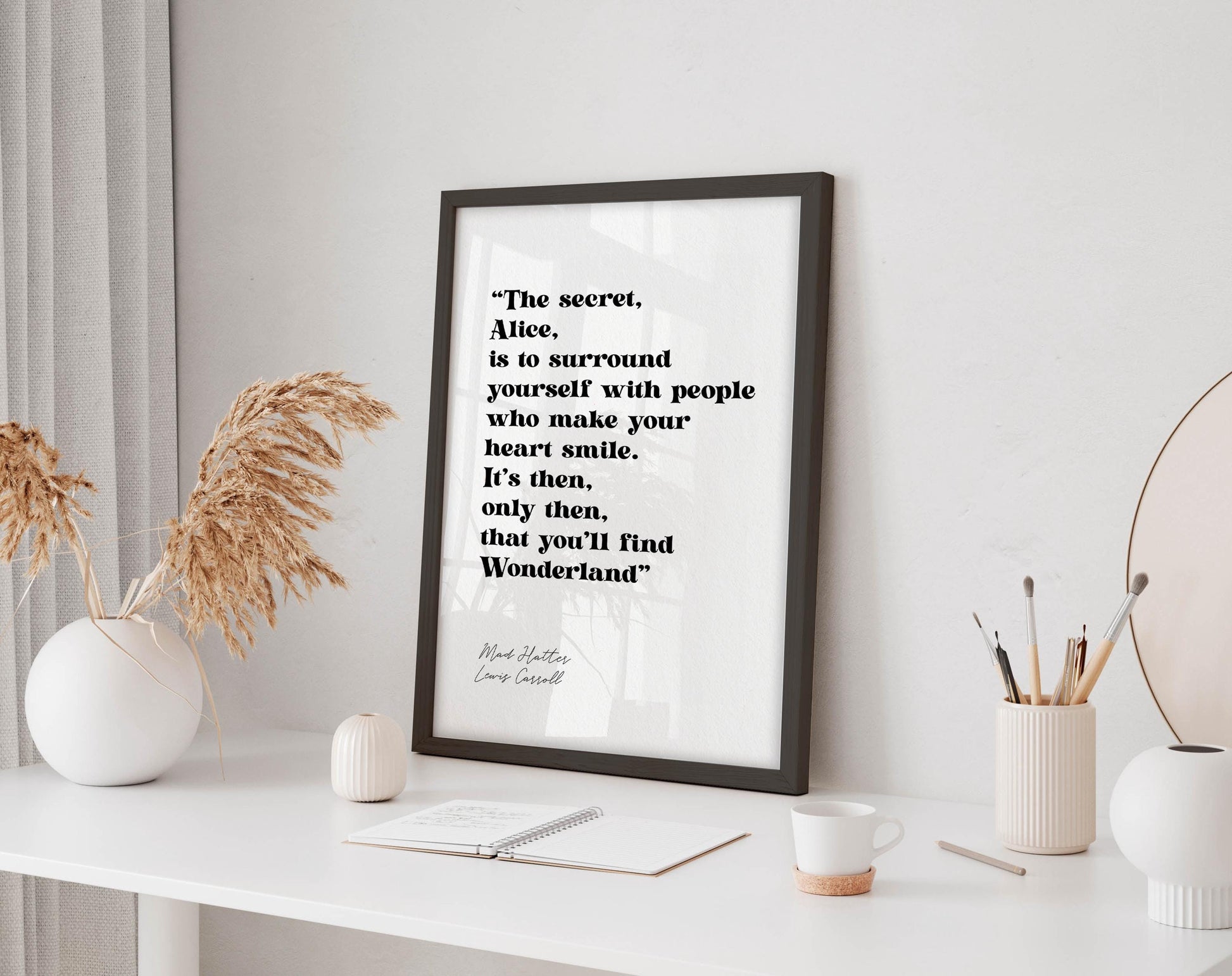 a framed quote by Lewis Carroll, &quot;The secret, Alice, is to surround yourself with people who make your heart smile. It&#39;s then, only you&#39;ll find Wonderland.&quot; The quote is displayed on a white wall, accompanied by a small notebook, a white mug, and a white vase with dried flowers.