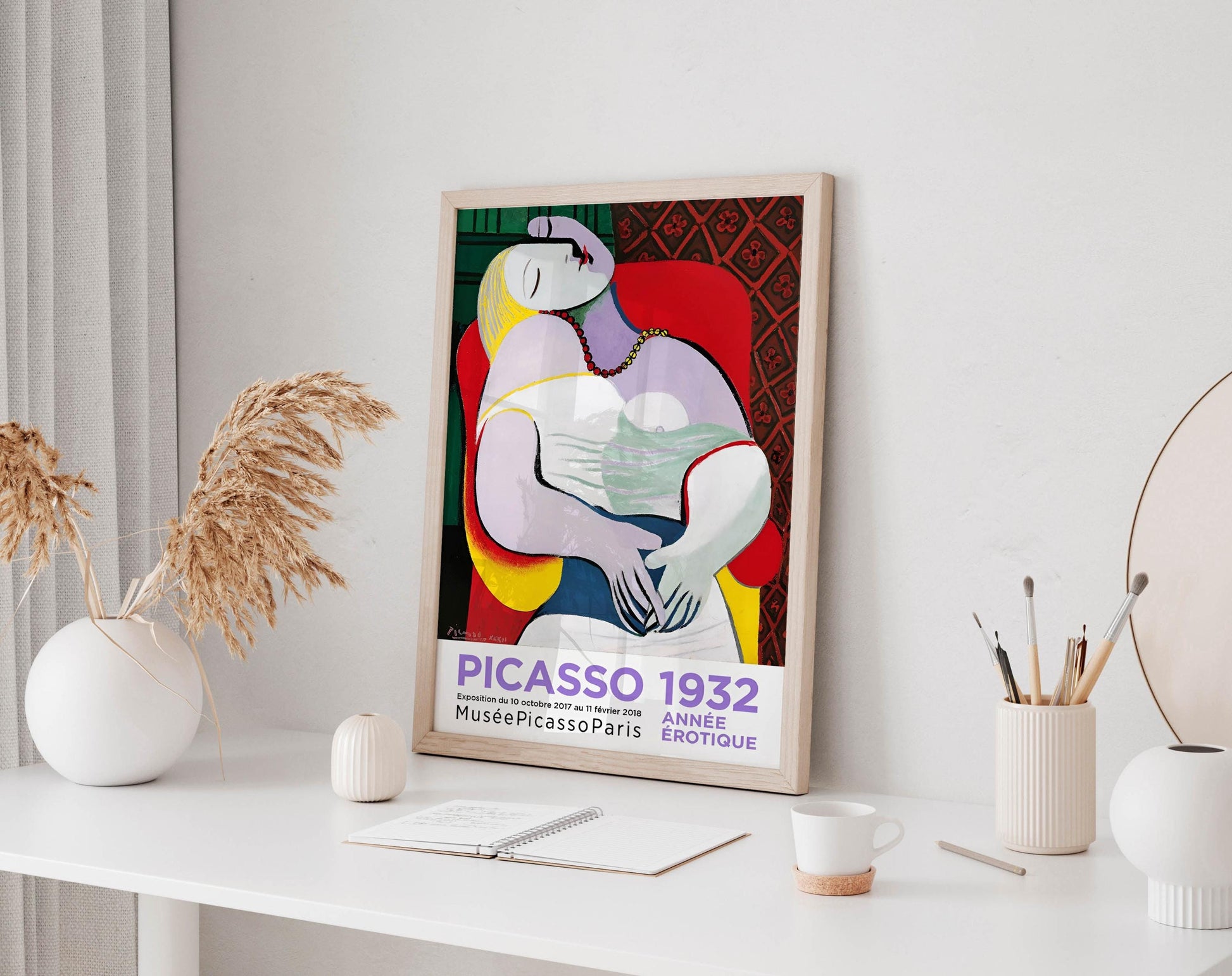 a framed poster of Picasso&#39;s cubist artwork, which is displayed on a white desk. The poster is surrounded by various objects, including a white vase, a small white candle, a white mug, and a white notebook.