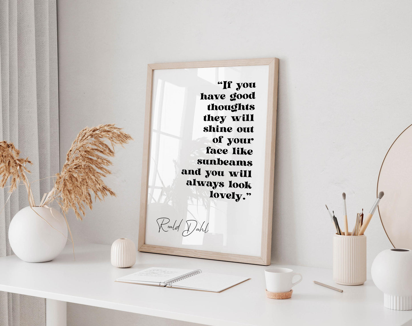 a framed quote by Ralph Dhl on a white wall, accompanied by a white desk with various items such as a notebook, a cup, and a vase with dried flowers.