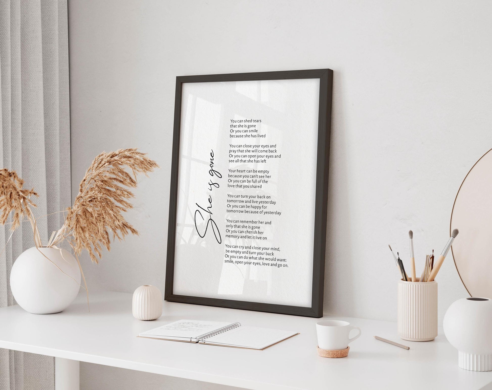 a framed poem titled &quot;She&#39;s gone&quot; displayed on a white desk, accompanied by a notebook, a cup, and a vase with dried flowers.