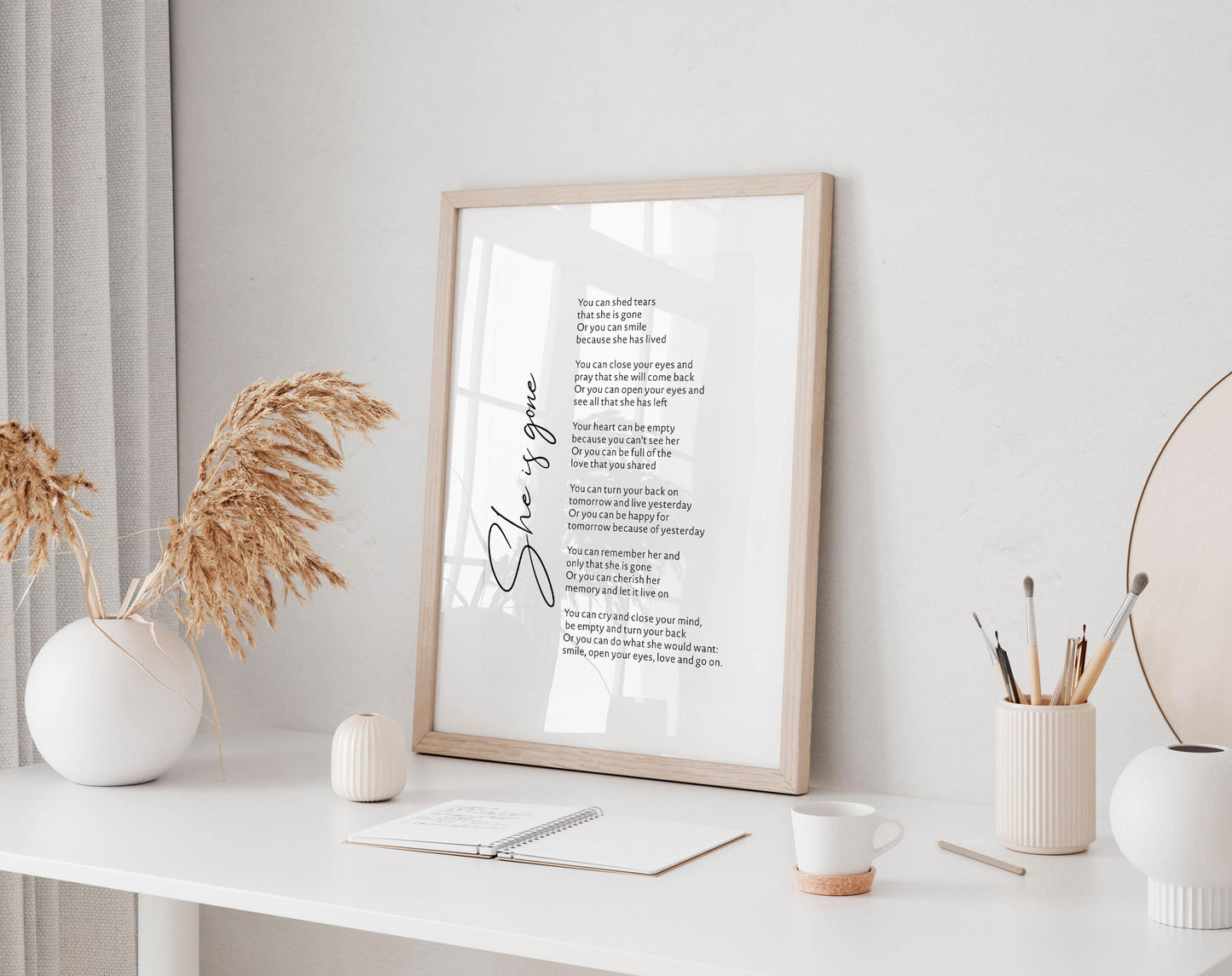 a framed poem titled &quot;She&#39;s gone&quot; displayed on a white desk, accompanied by a notebook, a cup, and a vase with dried flowers.