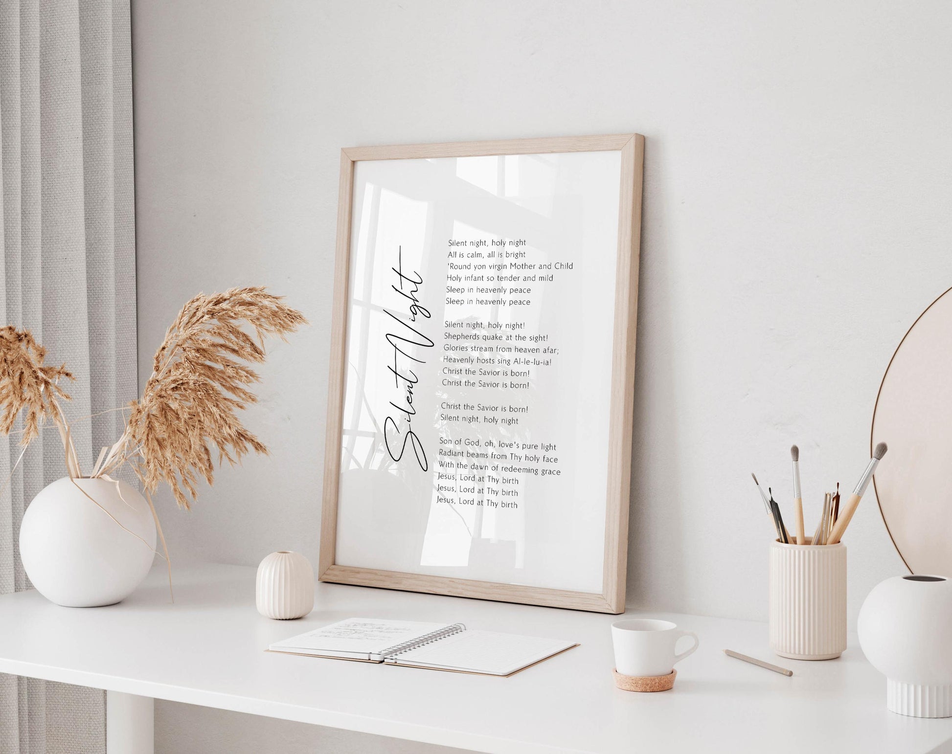 a framed poem titled &quot;Silent Night&quot; on a white desk, with a notebook, a cup, and a vase of dried flowers nearby.