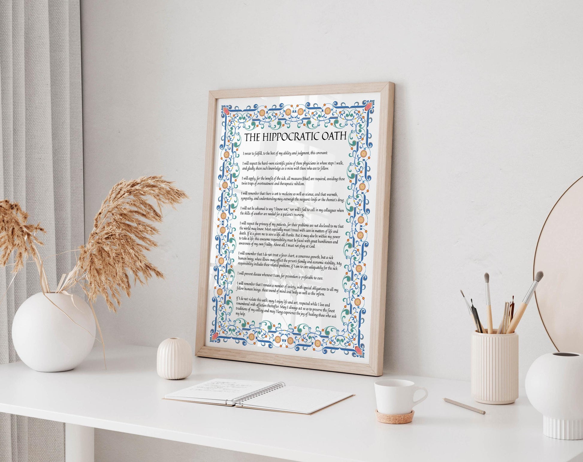 a framed poster of a text on a white wall, with a white desk in the foreground holding a notebook, a cup, and a vase with dried flowers.