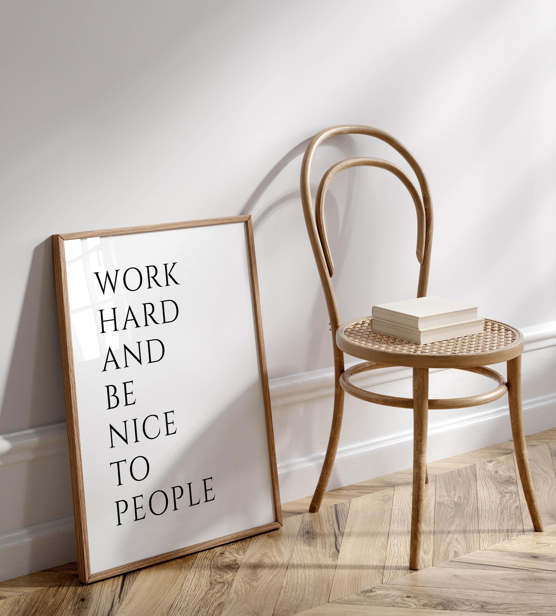 a wooden chair with a book on it, placed next to a framed poster with the text &quot;WORK HARD AND BE NICE TO PEOPLE&quot; on a white wall.