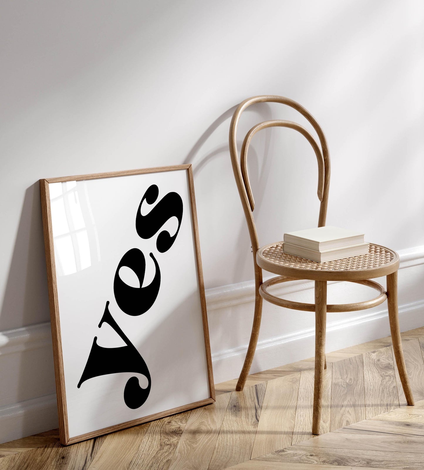 a wooden chair with a stack of books on it, placed against a white wall. There is a framed poster with the word &quot;Yes&quot; written on it, positioned on the floor next to the chair.