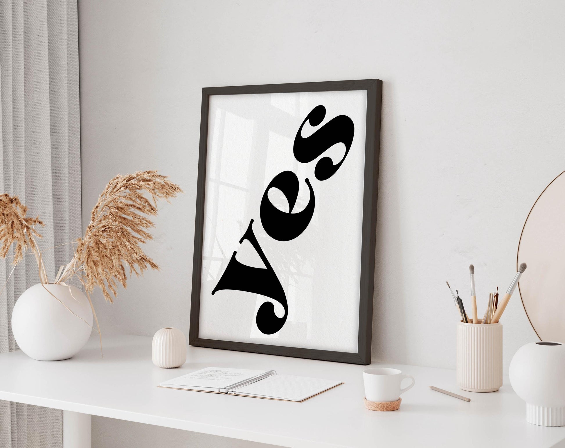 a white desk with a framed poster of the word &quot;Yes&quot; on it, along with a notebook, a cup, and some decorative elements such as a vase and a potted plant.