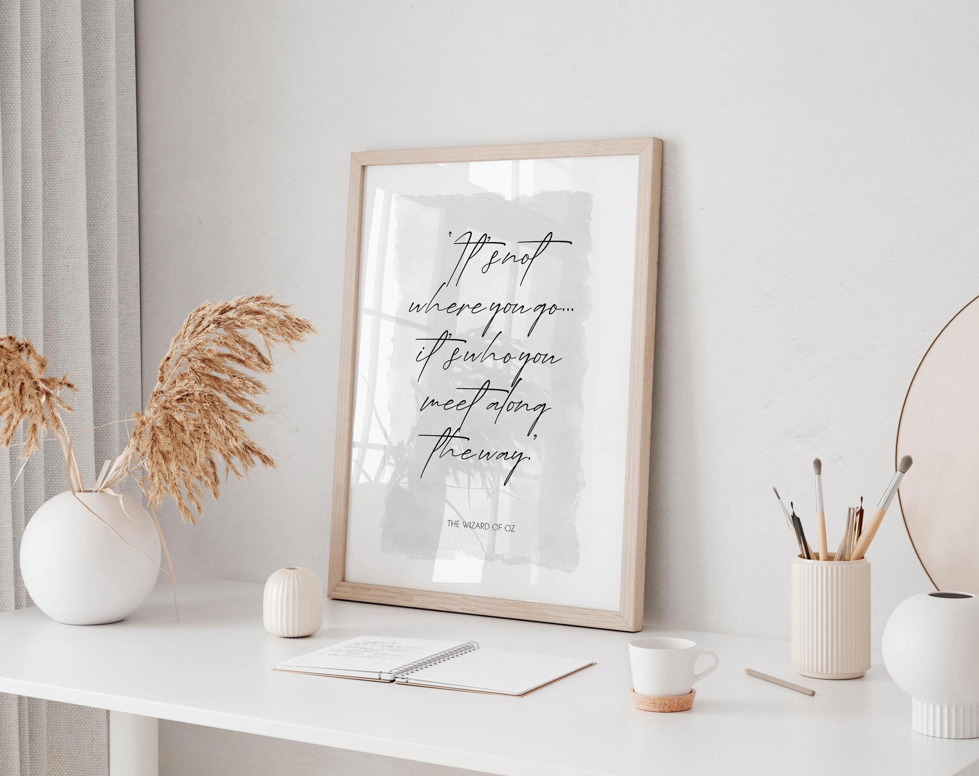 a framed quote on a white desk, surrounded by various objects such as a notebook, a cup, and a vase with dried flowers.