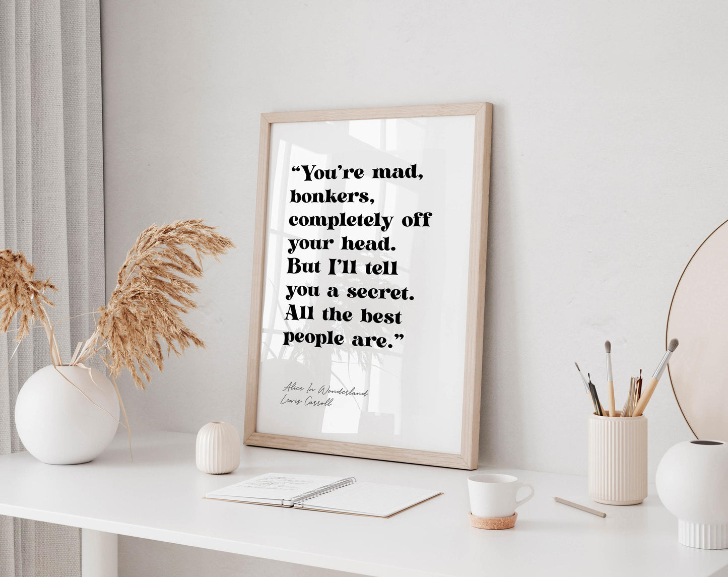 a framed quote on a white wall, surrounded by various objects such as a notebook, a cup, and a vase with dried flowers.