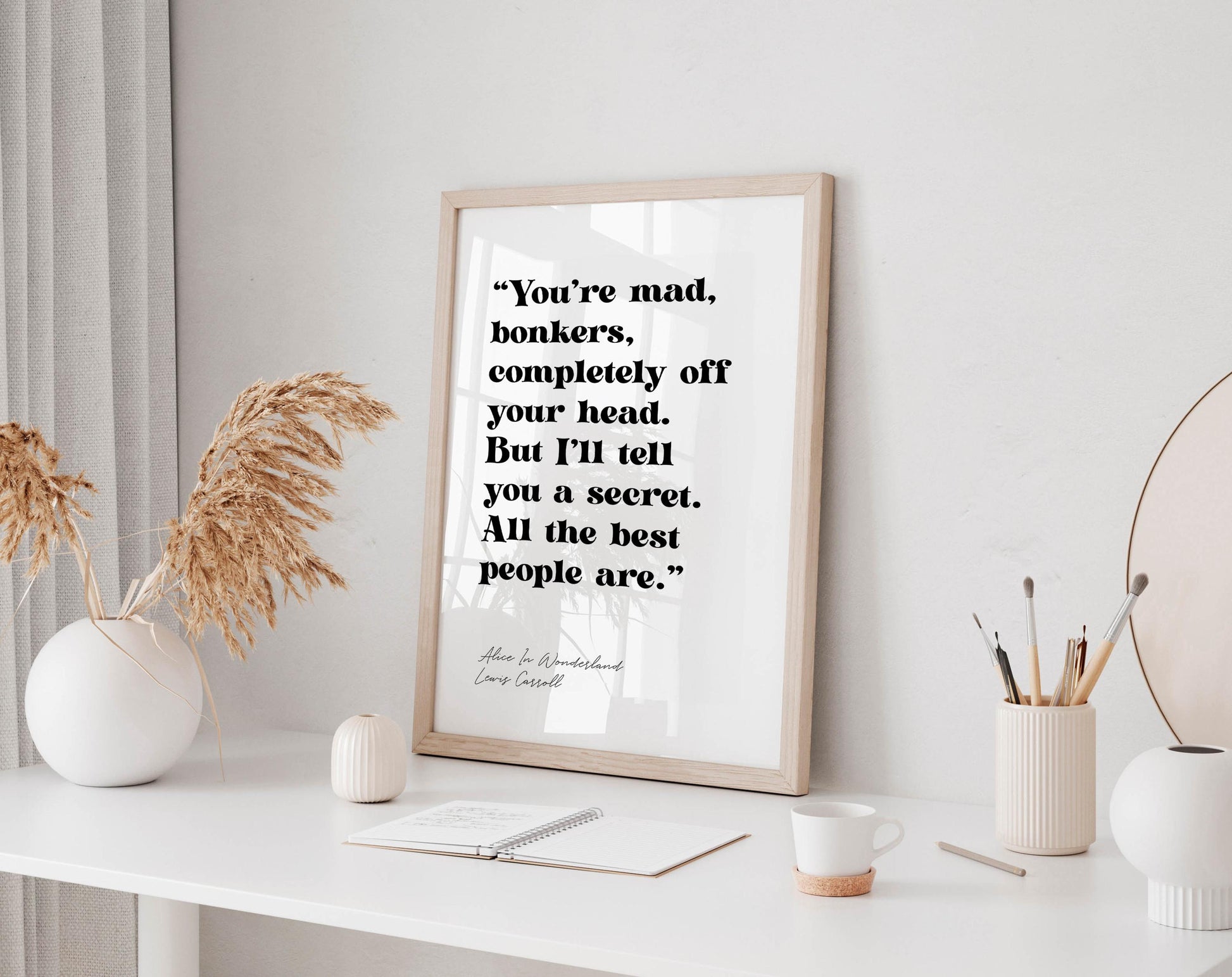 a framed quote on a white wall, surrounded by various objects such as a notebook, a cup, and a vase with dried flowers.