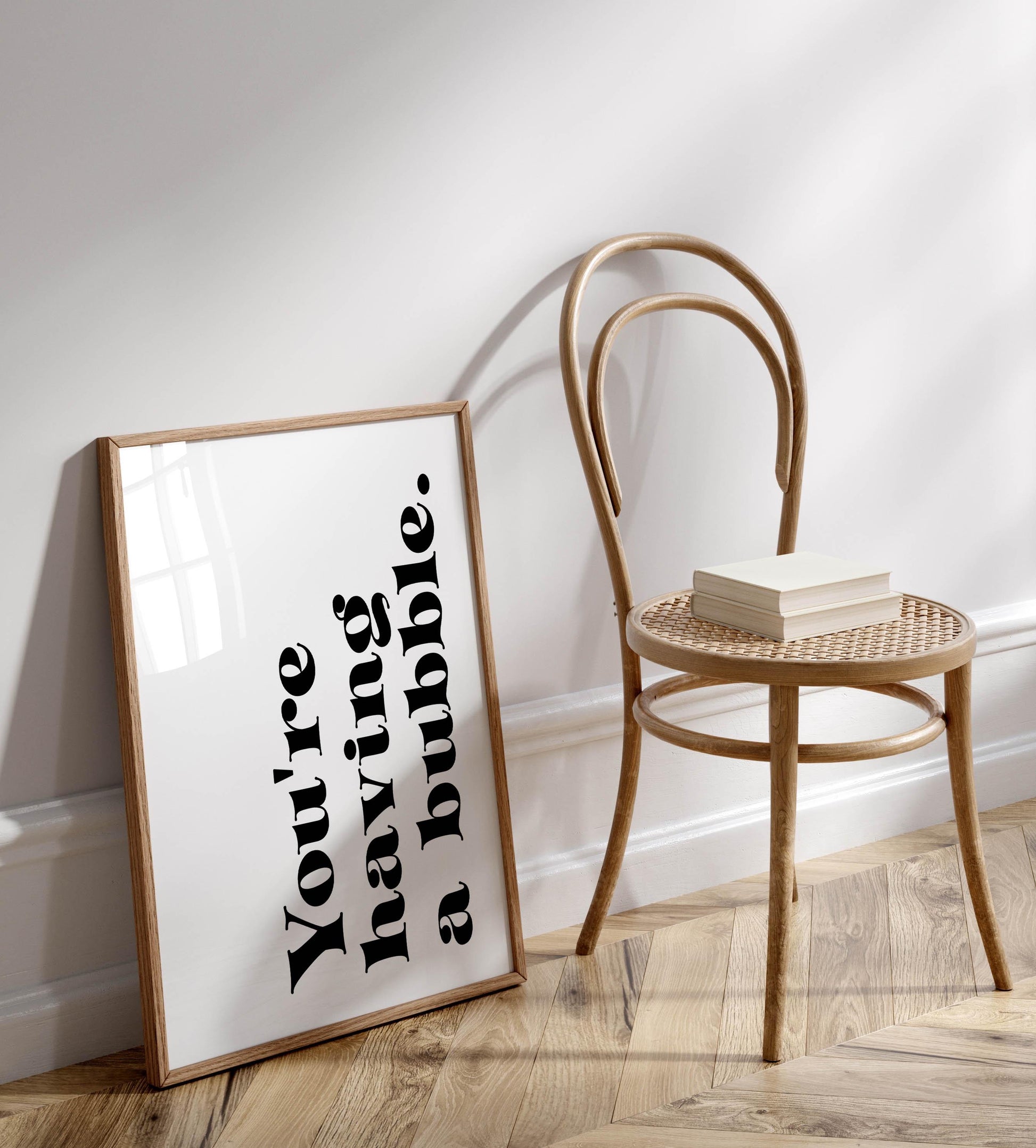 a white wall with a framed quote that reads &quot;You&#39;re having a bubble&quot; in black text. Next to the wall is a wooden chair with a stack of books on it.