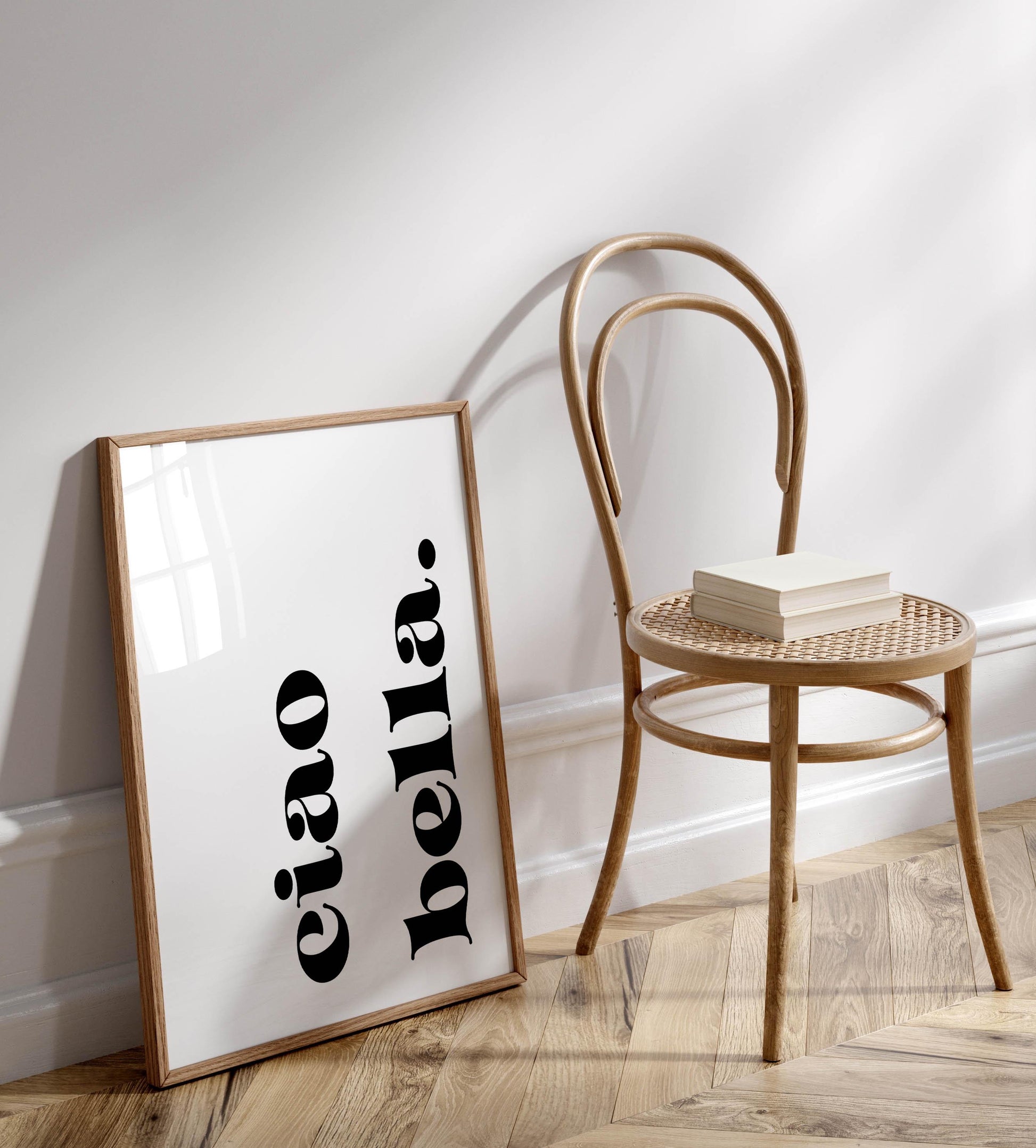 a wooden chair with a book on it, placed next to a framed poster with the text &quot;eiao bella&quot; on it, which is leaning against a white wall.