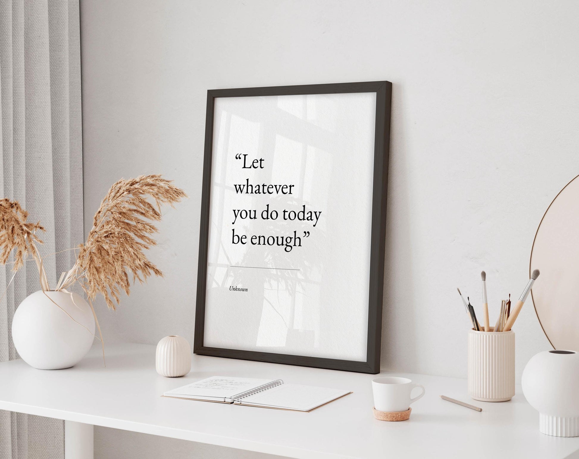 a framed quote on a white desk, surrounded by various objects such as a notebook, a cup, and a vase with dried flowers.
