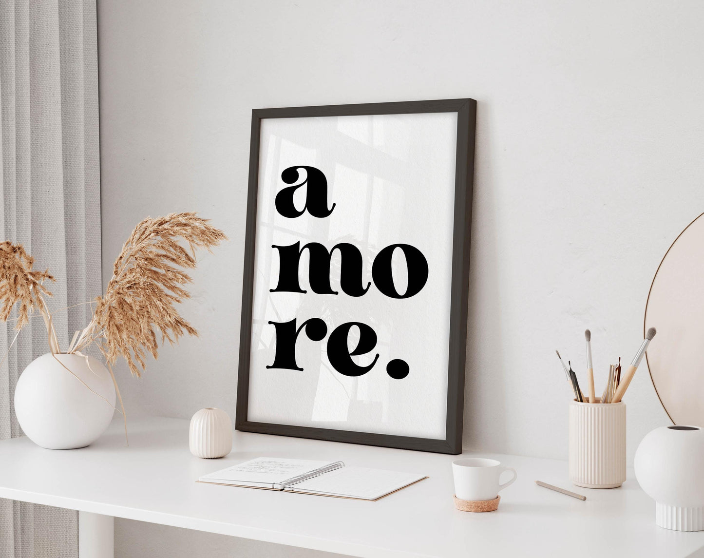 a white desk with a framed poster displaying the text &quot;a more.&quot; The desk also has various items such as a cup, a notebook, and a vase with dried flowers.