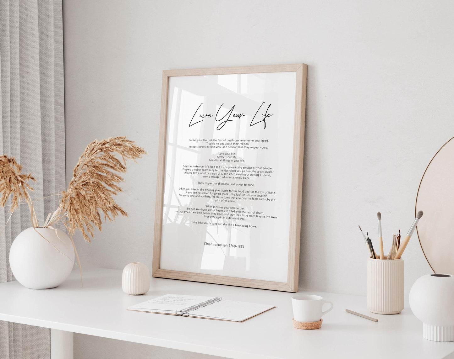 a framed inspirational quote, a white desk with various items such as a notebook, a cup, and a vase with dried flowers, all set against a white wall.