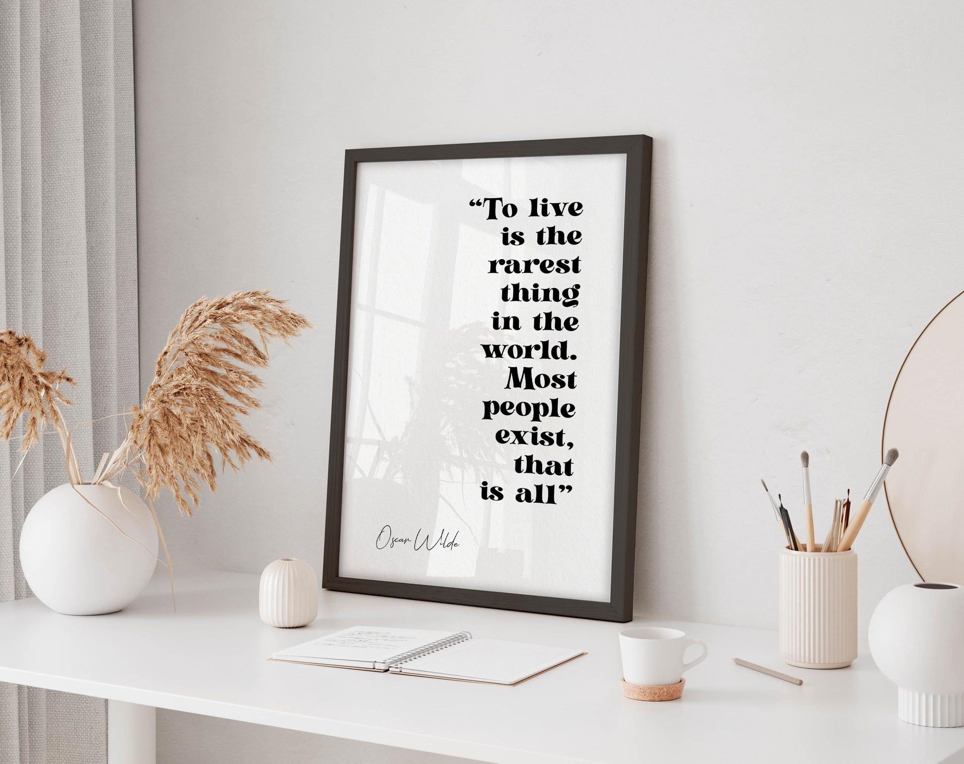 a framed quote by Oscar Wilde, &quot;To live is the rarest thing in the world. Most people exist, that is all&quot;, placed on a white desk with a notebook, pen, and a cup of coffee.