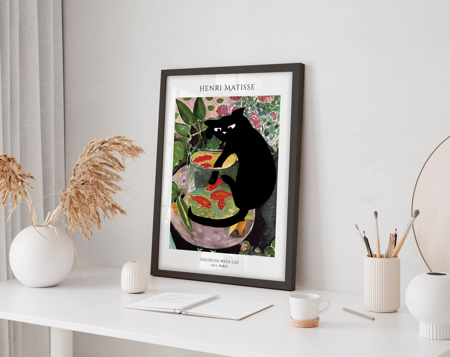 a framed painting of a black cat sitting in a fish bowl, surrounded by colorful fish and plants, placed on a white desk.
