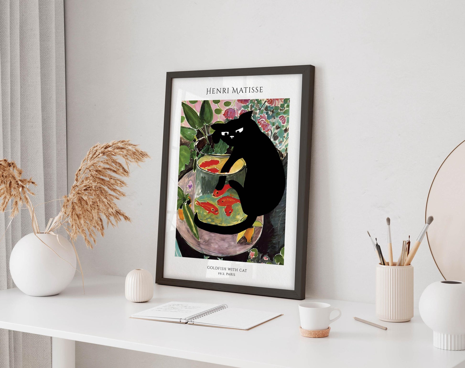 a framed painting of a black cat sitting in a fish bowl, surrounded by colorful fish and plants, placed on a white desk.