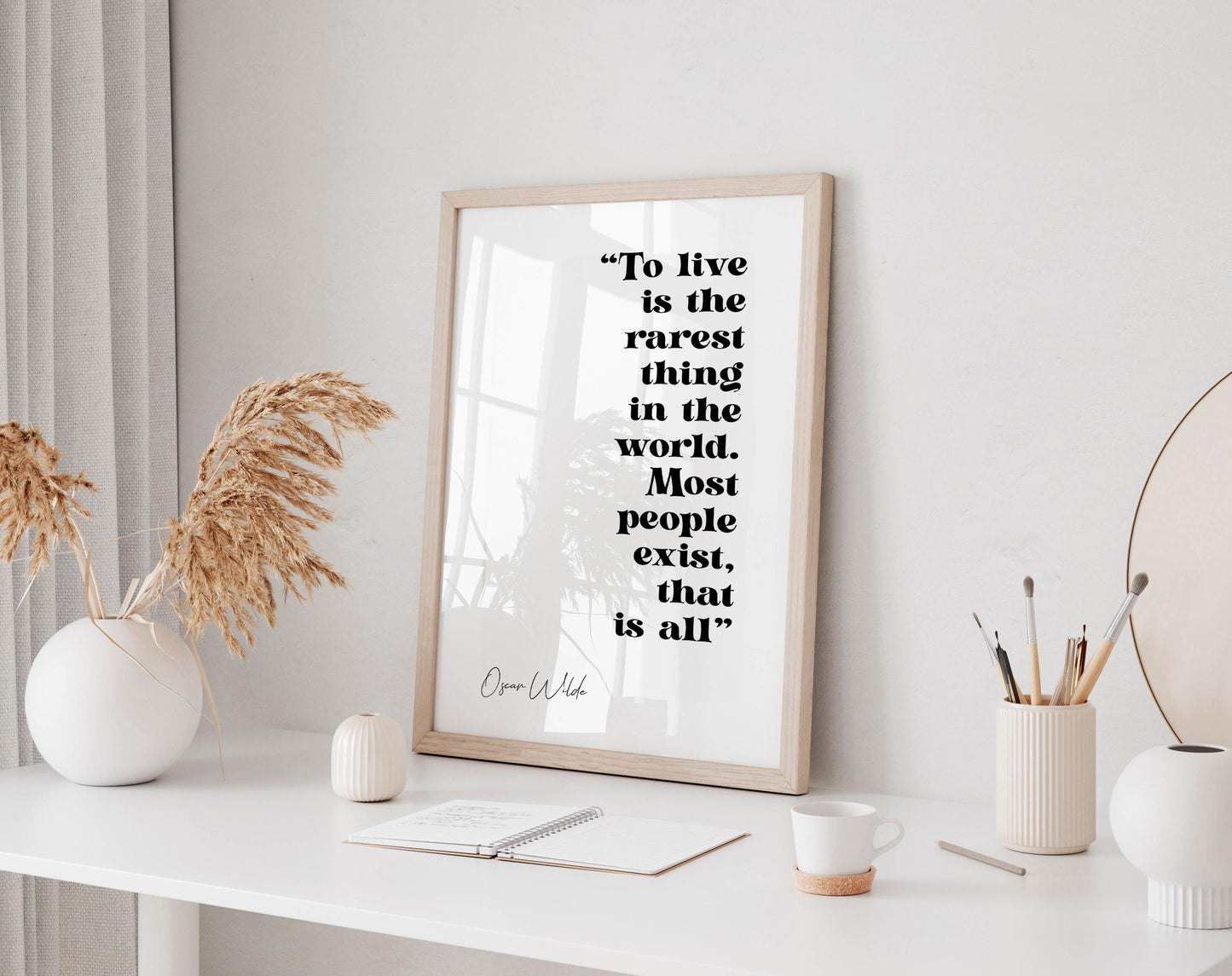a framed quote by Oscar Wilde, &quot;To live is the rarest thing in the world. Most people exist, that is all&quot;, displayed on a white wall above a desk with various items on it, including a vase with dried flowers, a notebook, a cup, and a pen holder.