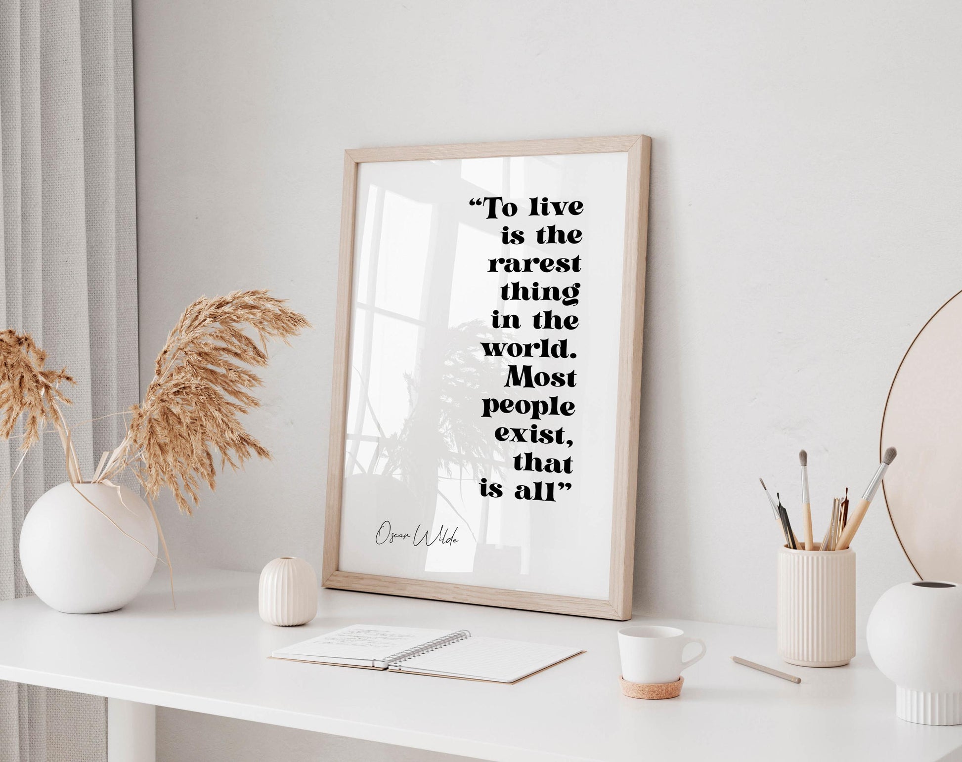 a framed quote by Oscar Wilde, &quot;To live is the rarest thing in the world. Most people exist, that is all&quot;, displayed on a white wall above a desk with various items on it, including a vase with dried flowers, a notebook, a cup, and a pen holder.