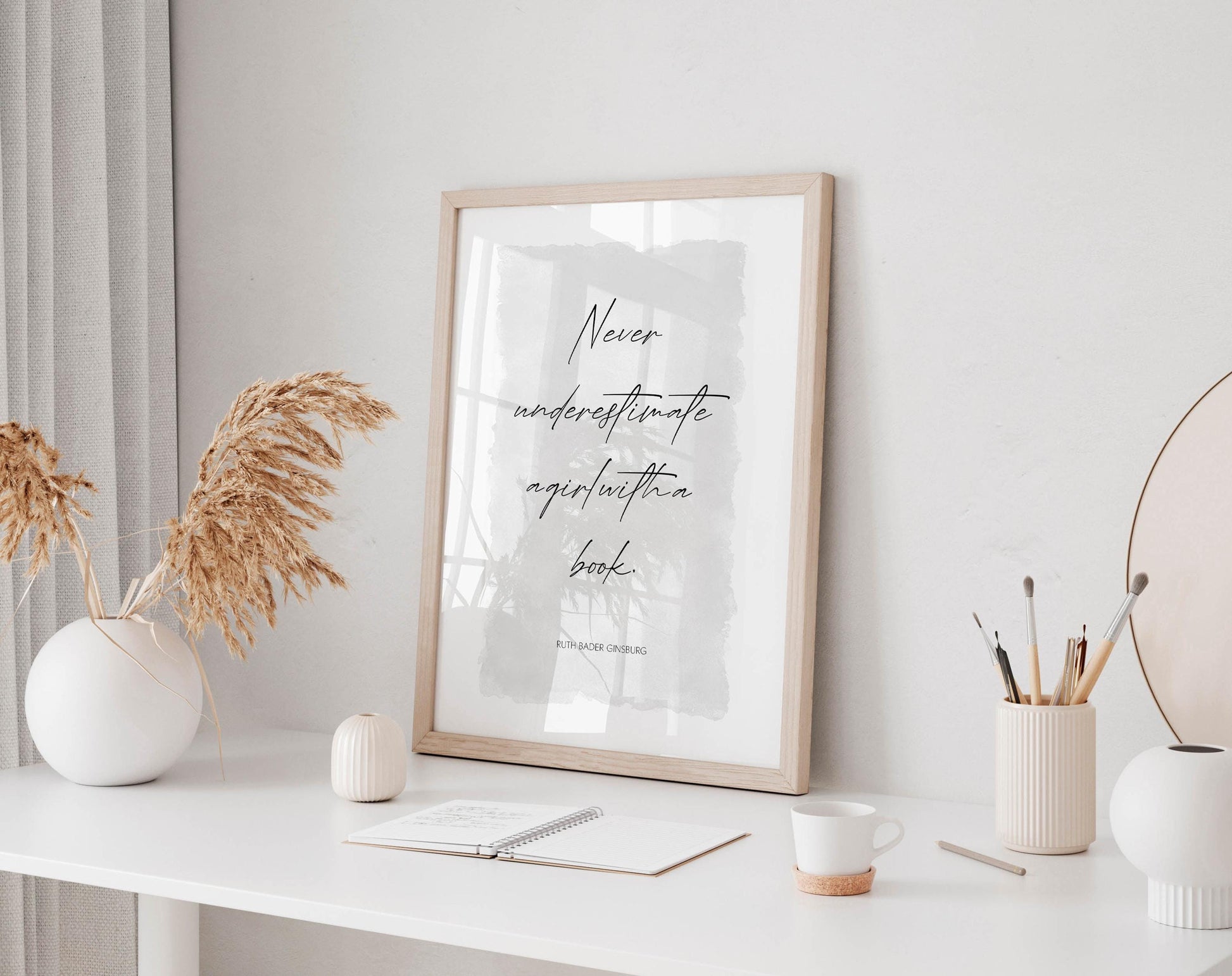 a minimalist workspace with a framed quote, a notebook, a cup, and a vase with dried flowers on a white desk.