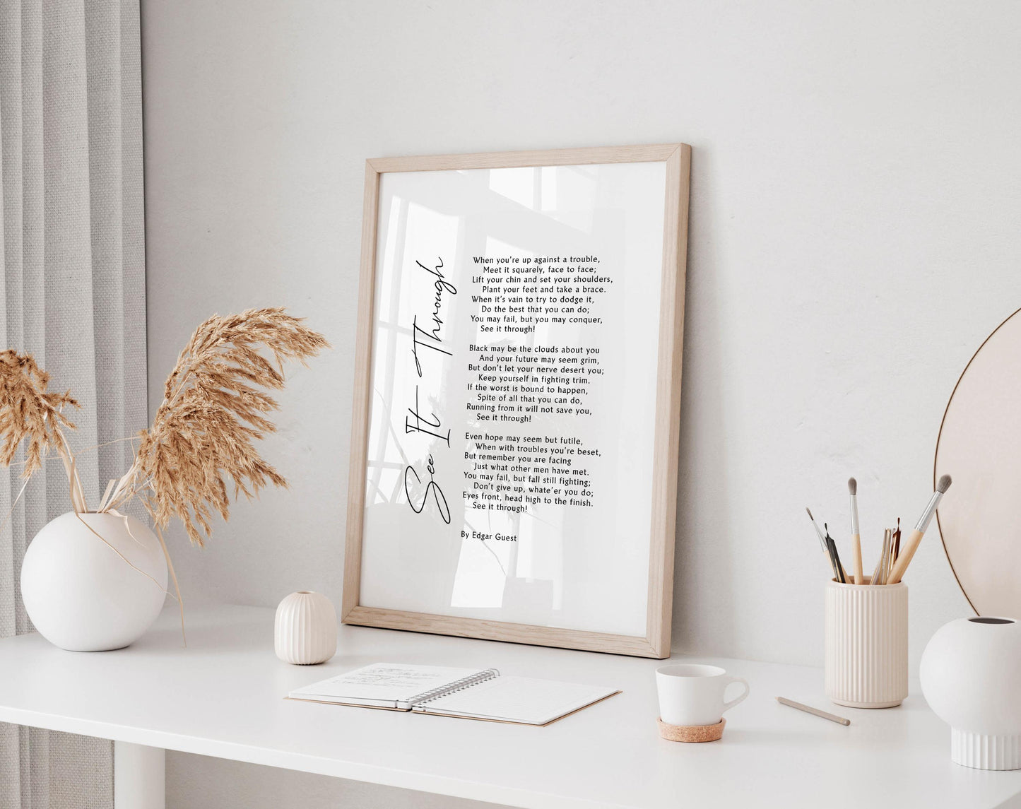 a framed poem titled &quot;See It Through&quot; displayed on a white desk, accompanied by a notebook, a cup, and a vase with dried flowers.