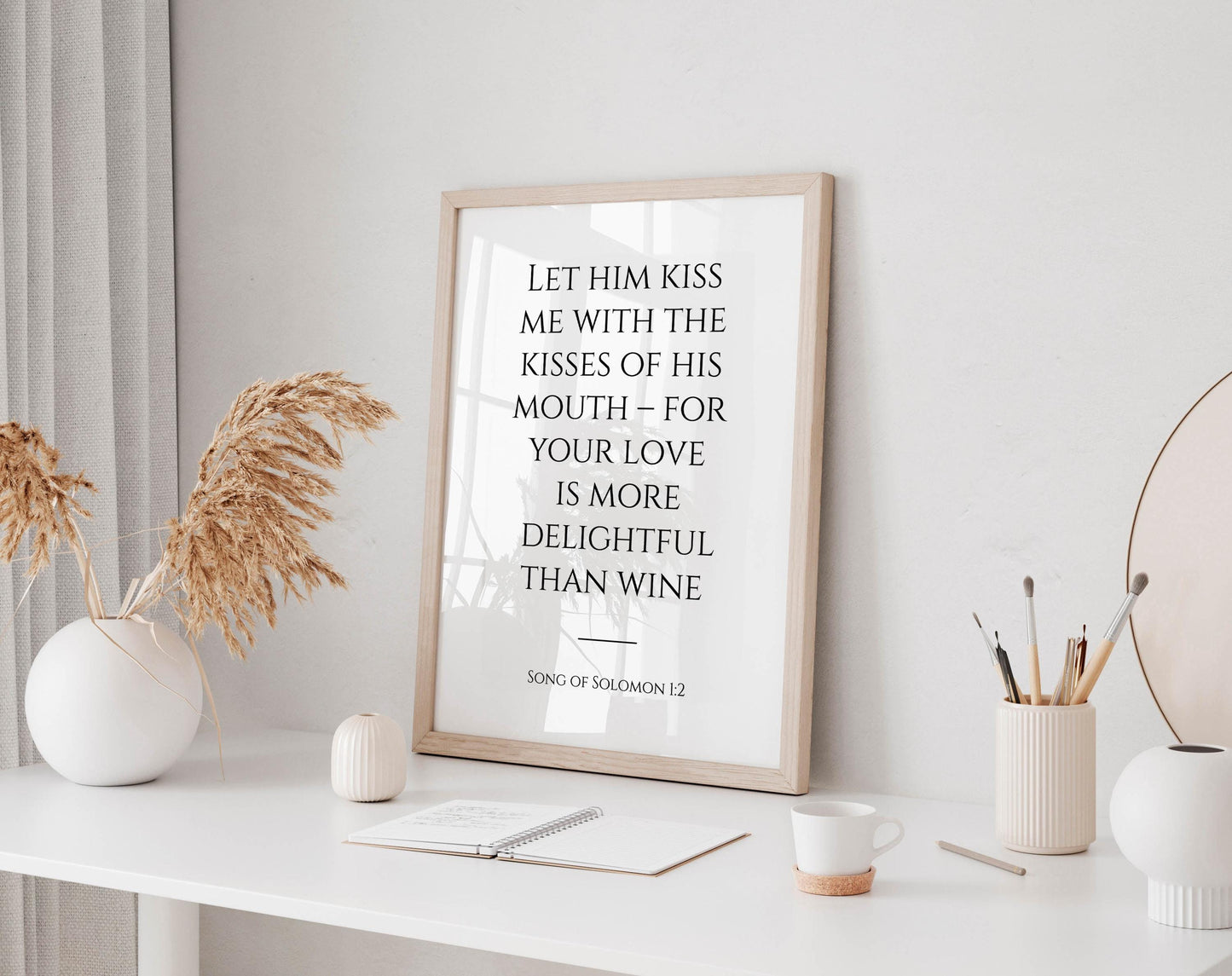 a framed quote on a white desk, surrounded by various objects such as a notebook, a cup, and a vase with dried flowers.