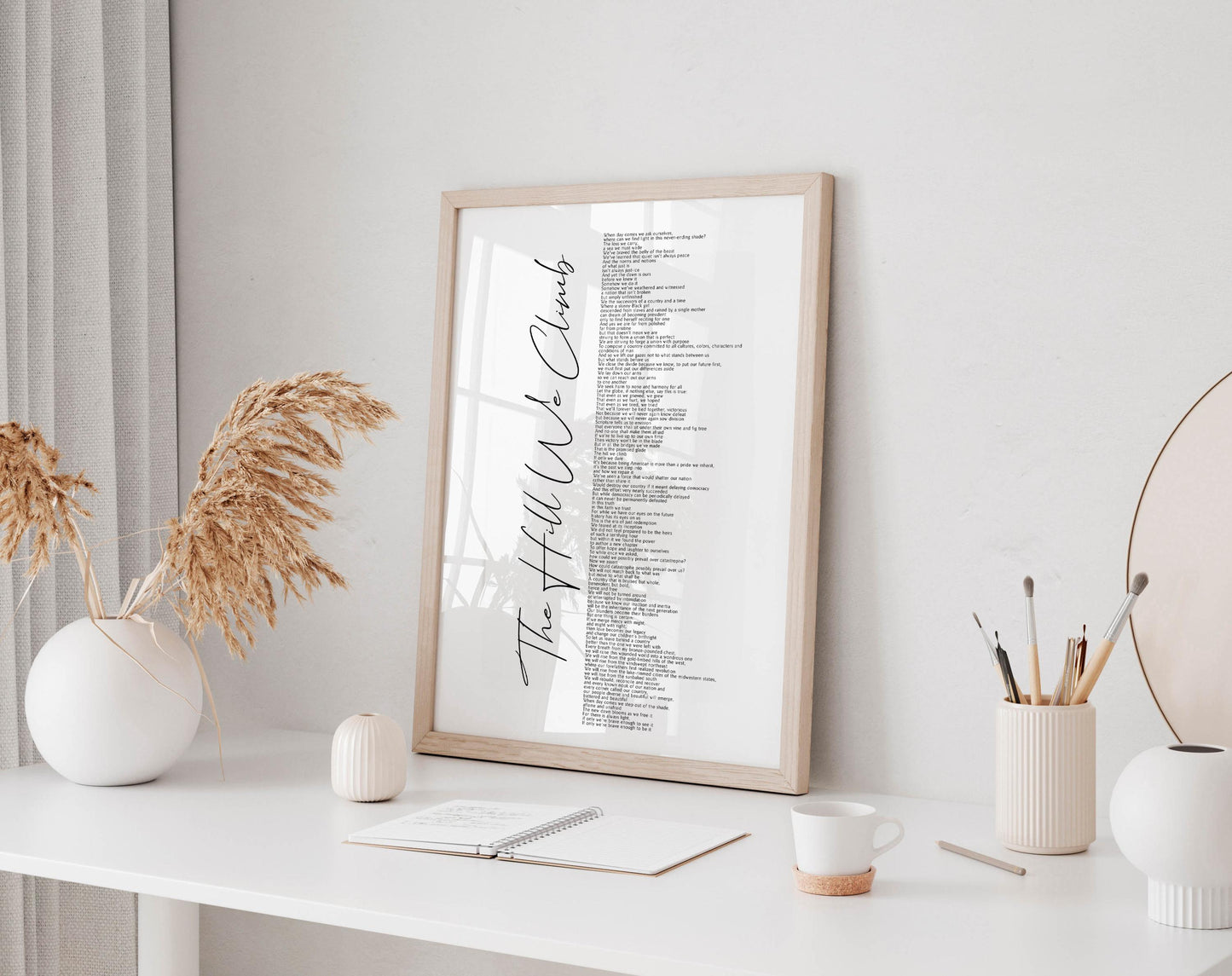 a white desk with a framed list of words, a notebook, a cup, and a vase with dried flowers.