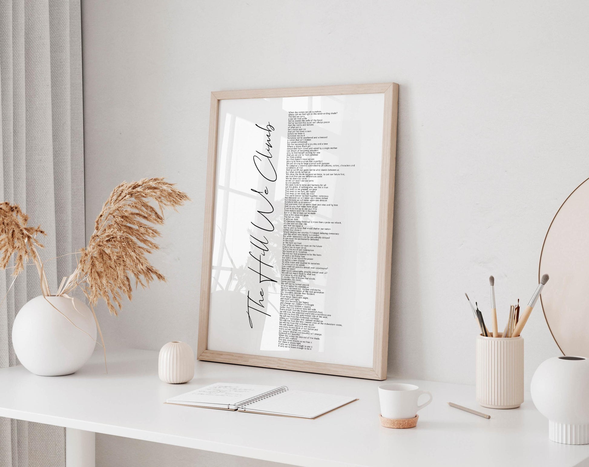 a white desk with a framed list of words, a notebook, a cup, and a vase with dried flowers.