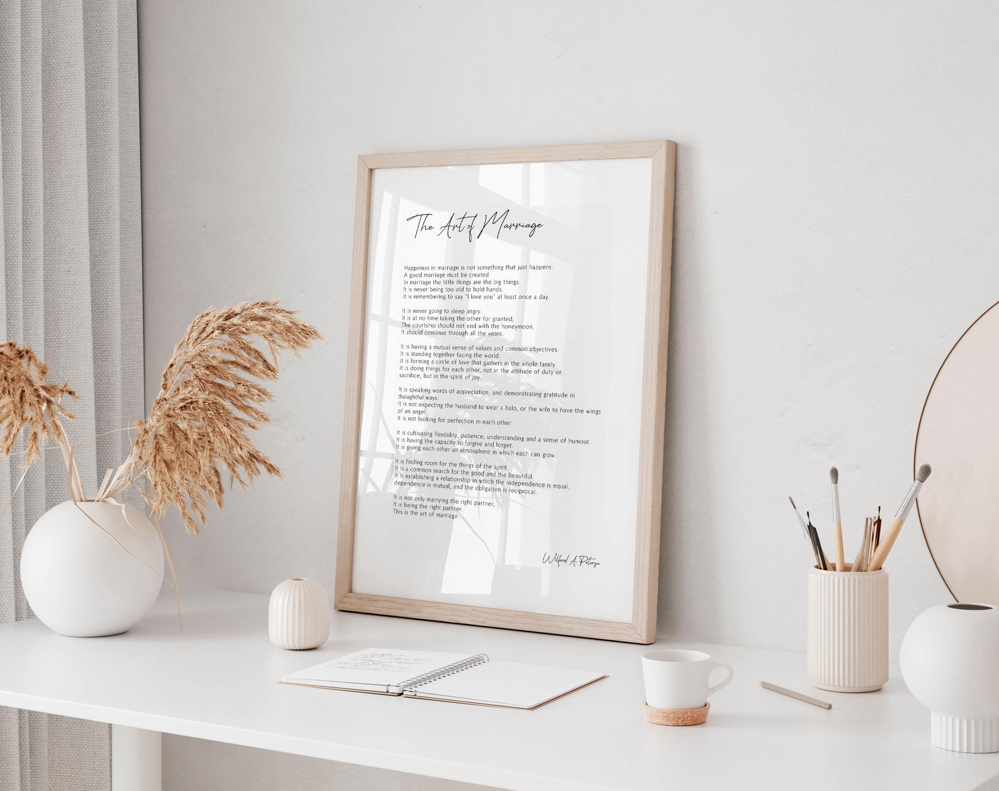 a framed poem titled &quot;The Art of Marriage&quot; displayed on a white desk, accompanied by a notebook, a cup, and a vase with dried flowers.