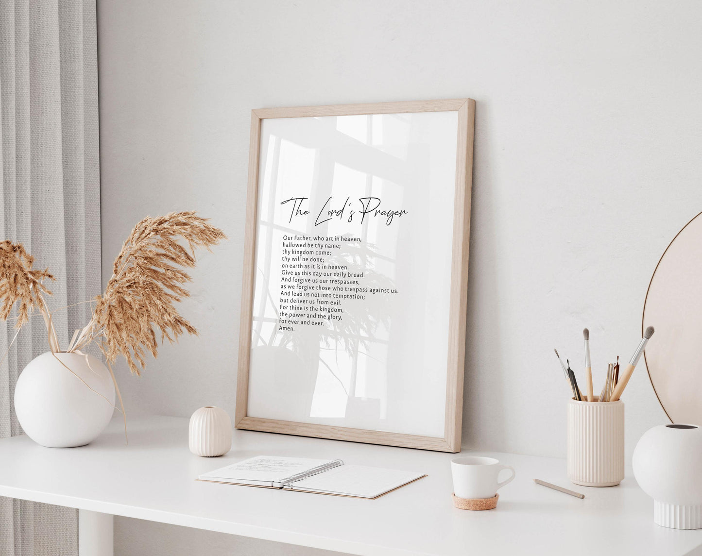 a white desk with a framed poster of a prayer, a white vase with dried flowers, a white mug, a notebook, and a small white candle.