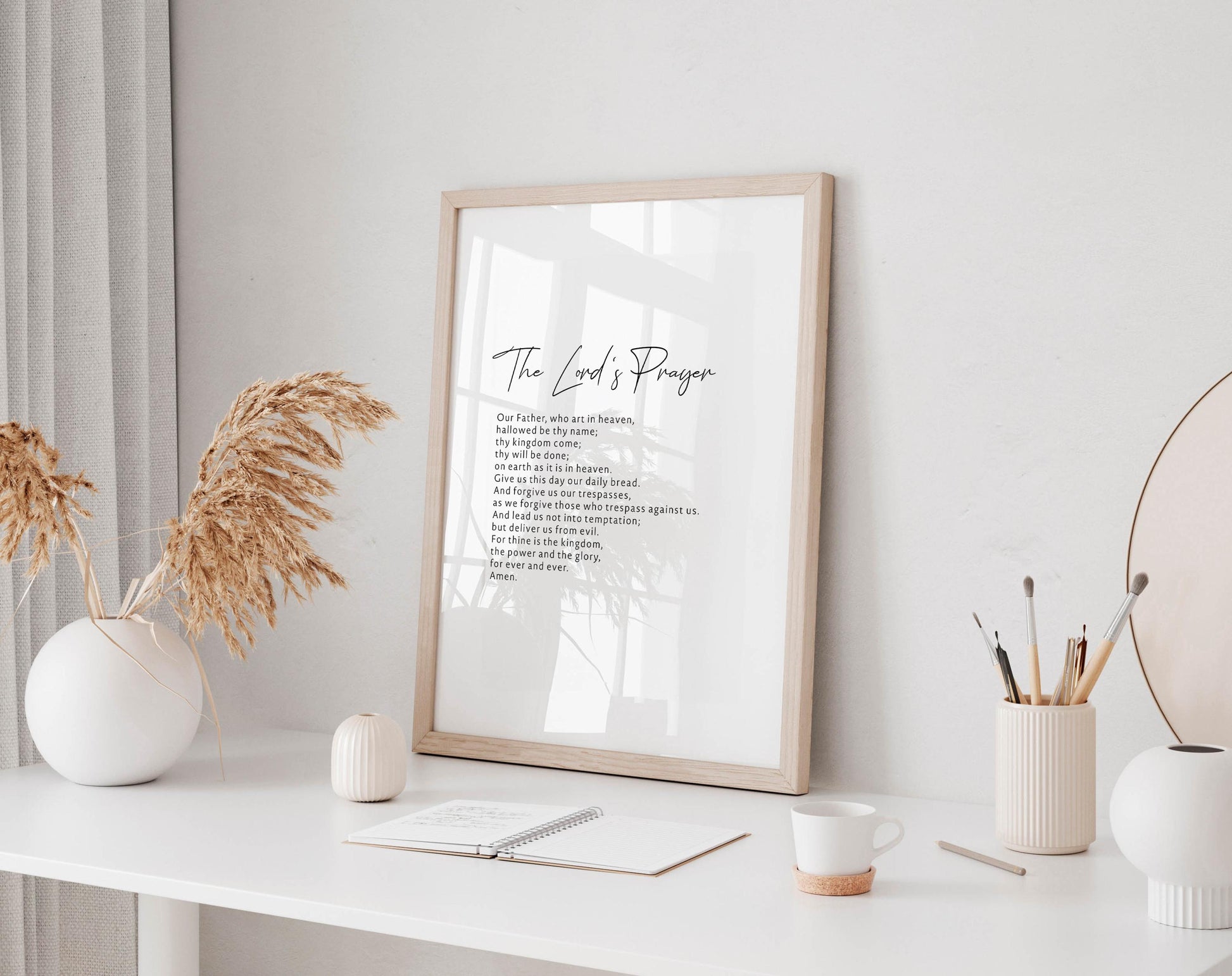 a white desk with a framed poster of a prayer, a white vase with dried flowers, a white mug, a notebook, and a small white candle.