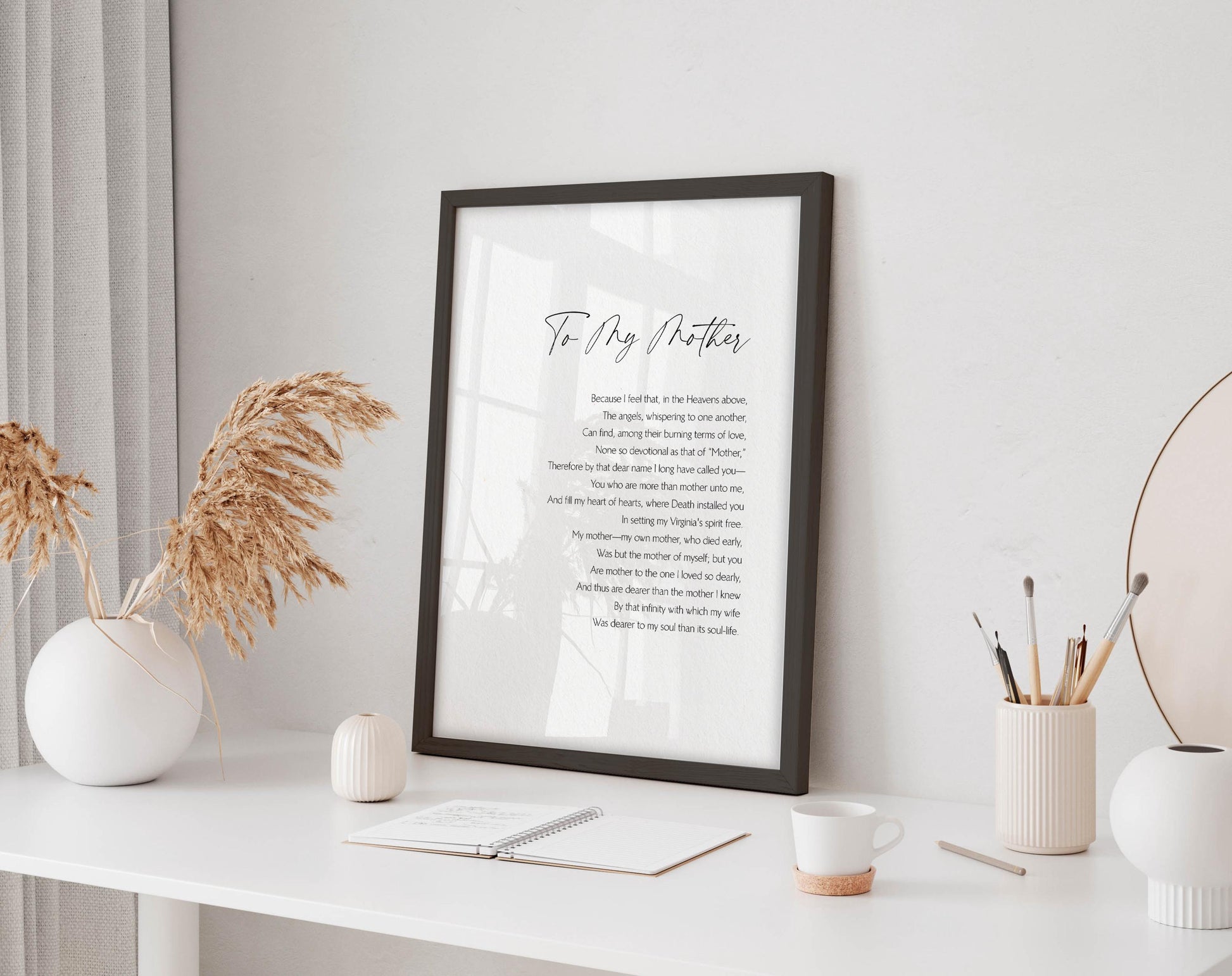 a framed poem titled &quot;To My Mother&quot; displayed on a white desk, accompanied by a notebook, a cup, and a vase with dried flowers.