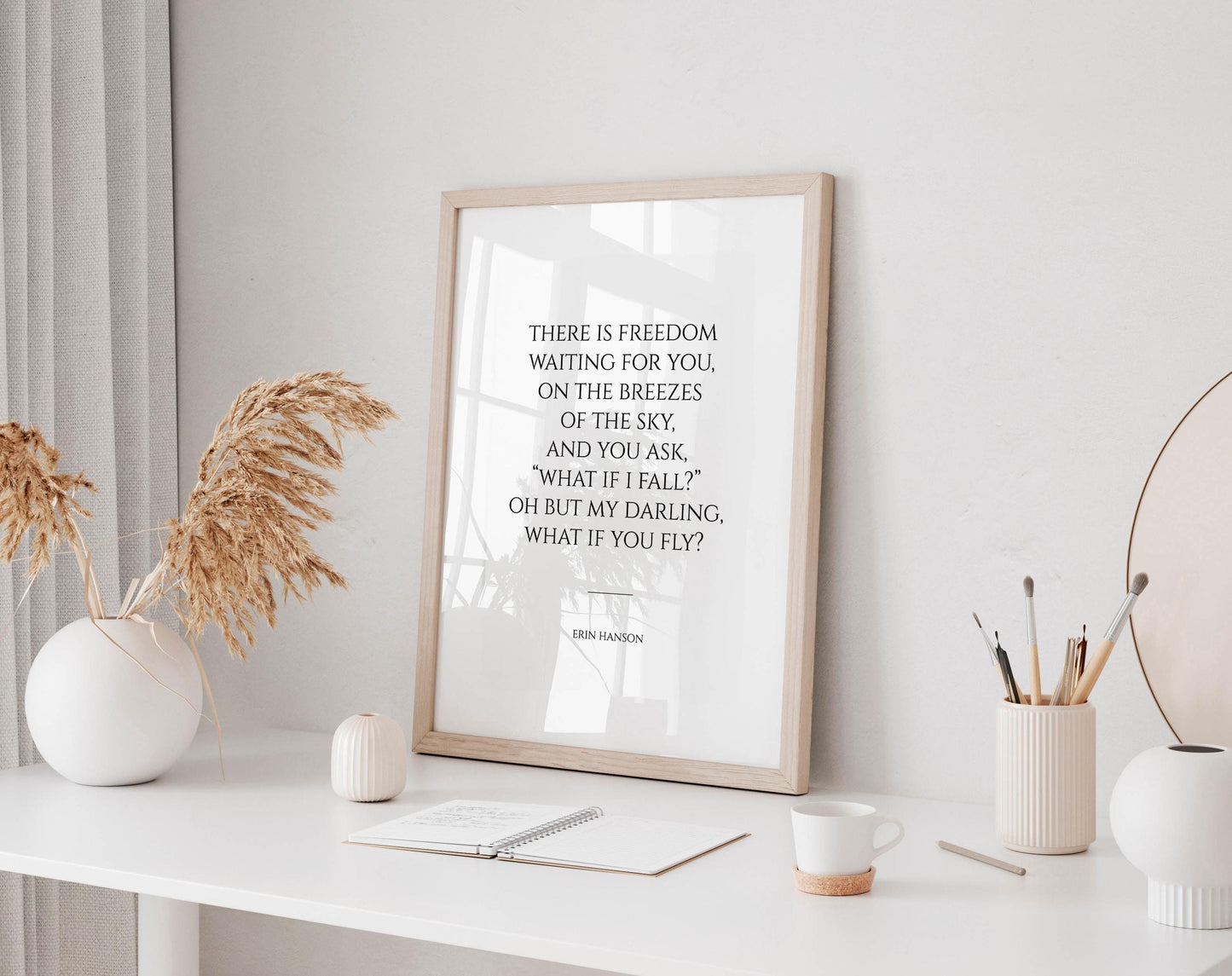 a framed quote on a white wall, accompanied by a desk with various items such as a notebook, a cup, and a vase with dried flowers.